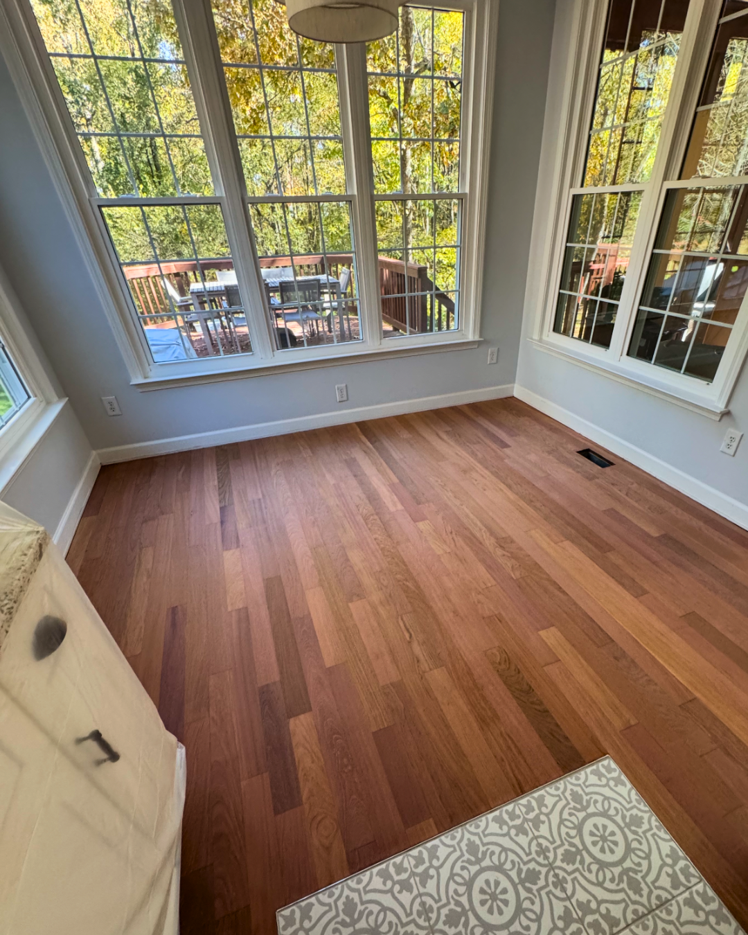 Empty room with hardwood flooring and large windows looking out onto a wooden deck with outdoor furniture, surrounded by trees.