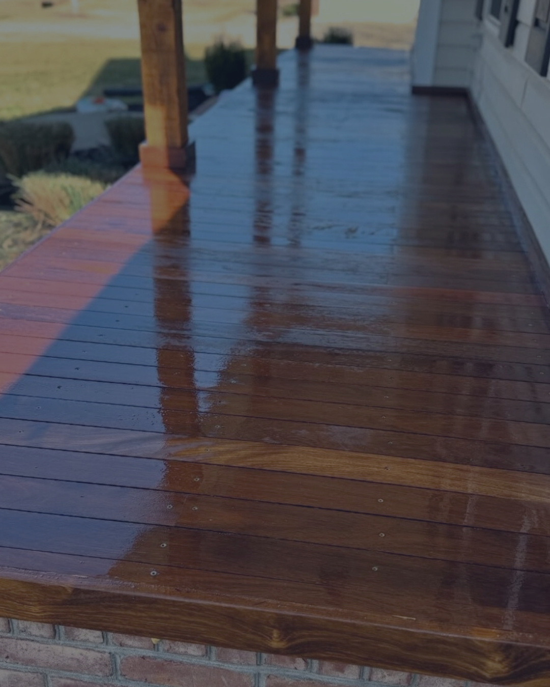 A recently cleaned and polished wooden deck on a house with a brick foundation, showing bright sunlight and shadows.