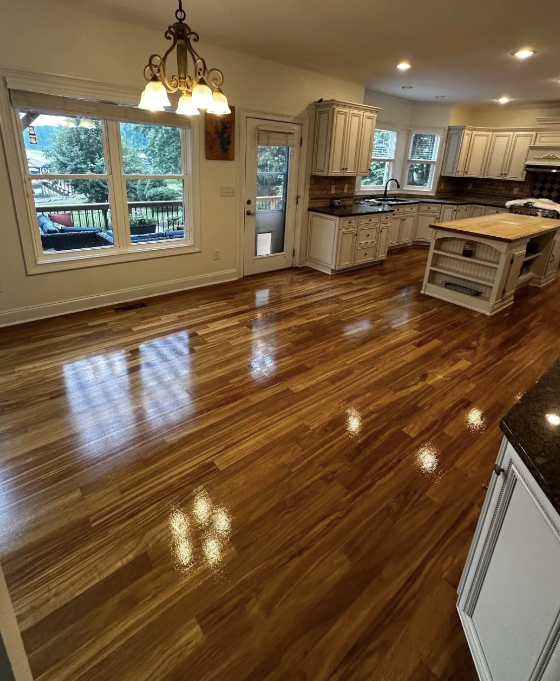 Empty kitchen and dining area with polished hardwood floors, cream-colored cabinets, a chandelier, large windows, and a view of an outdoor deck with seating.