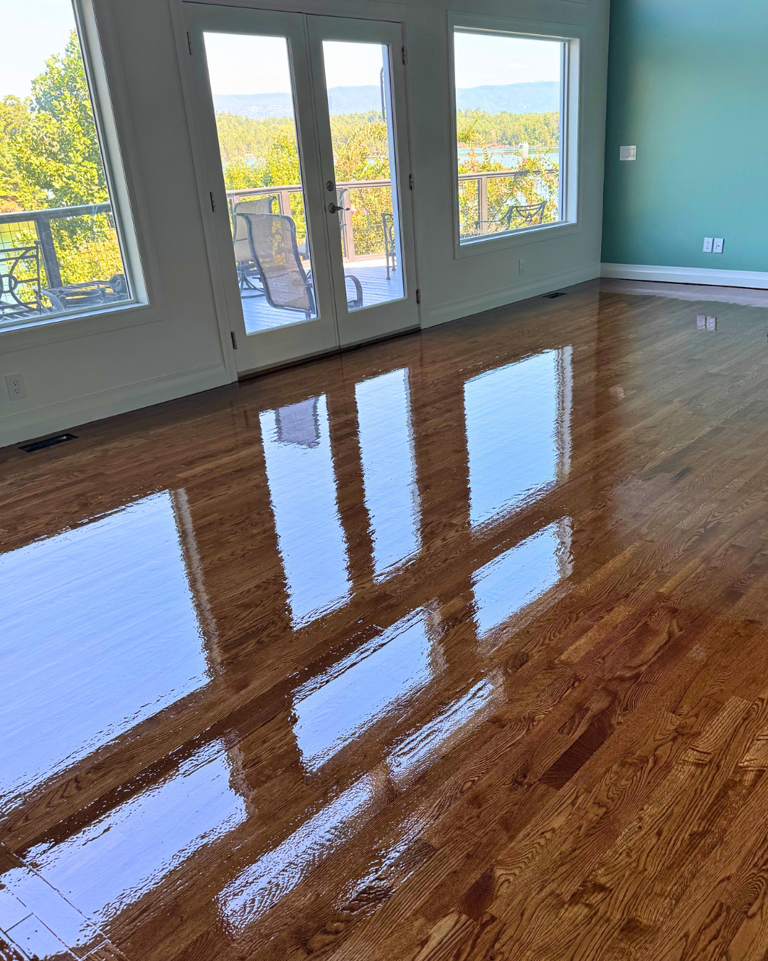 A room with a wooden floor that is freshly polished, reflecting windows, glass door, and outdoor scenery of trees and mountains.