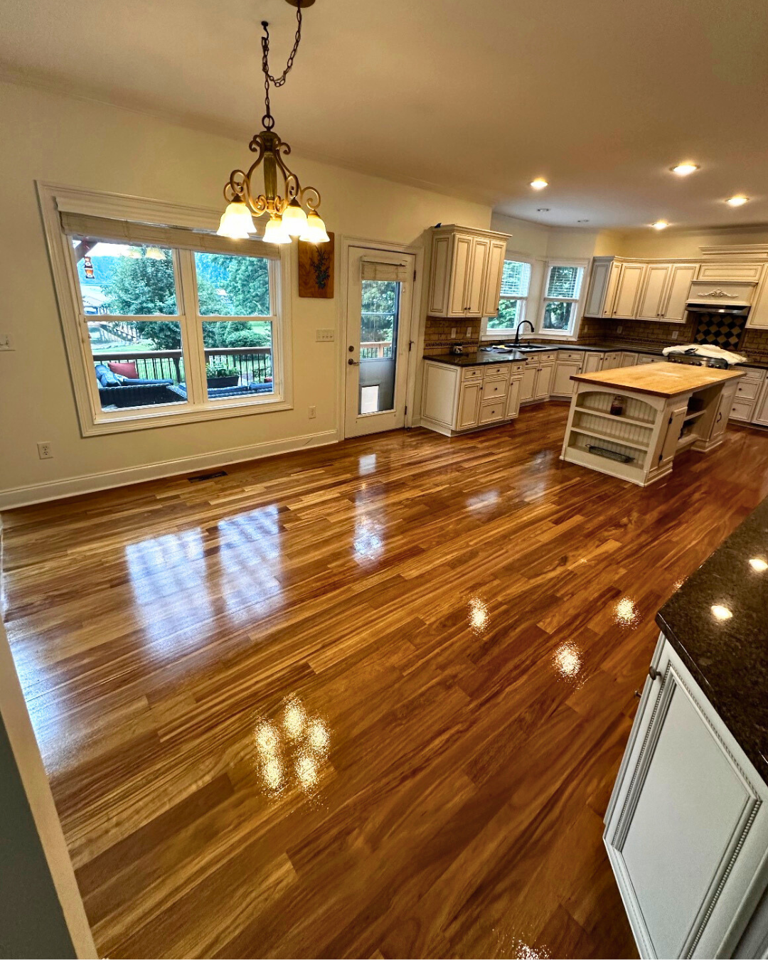 Empty kitchen and dining area with polished wooden floors, white cabinets, a kitchen island, windows overlooking a backyard, a chandelier, and recessed ceiling lights.