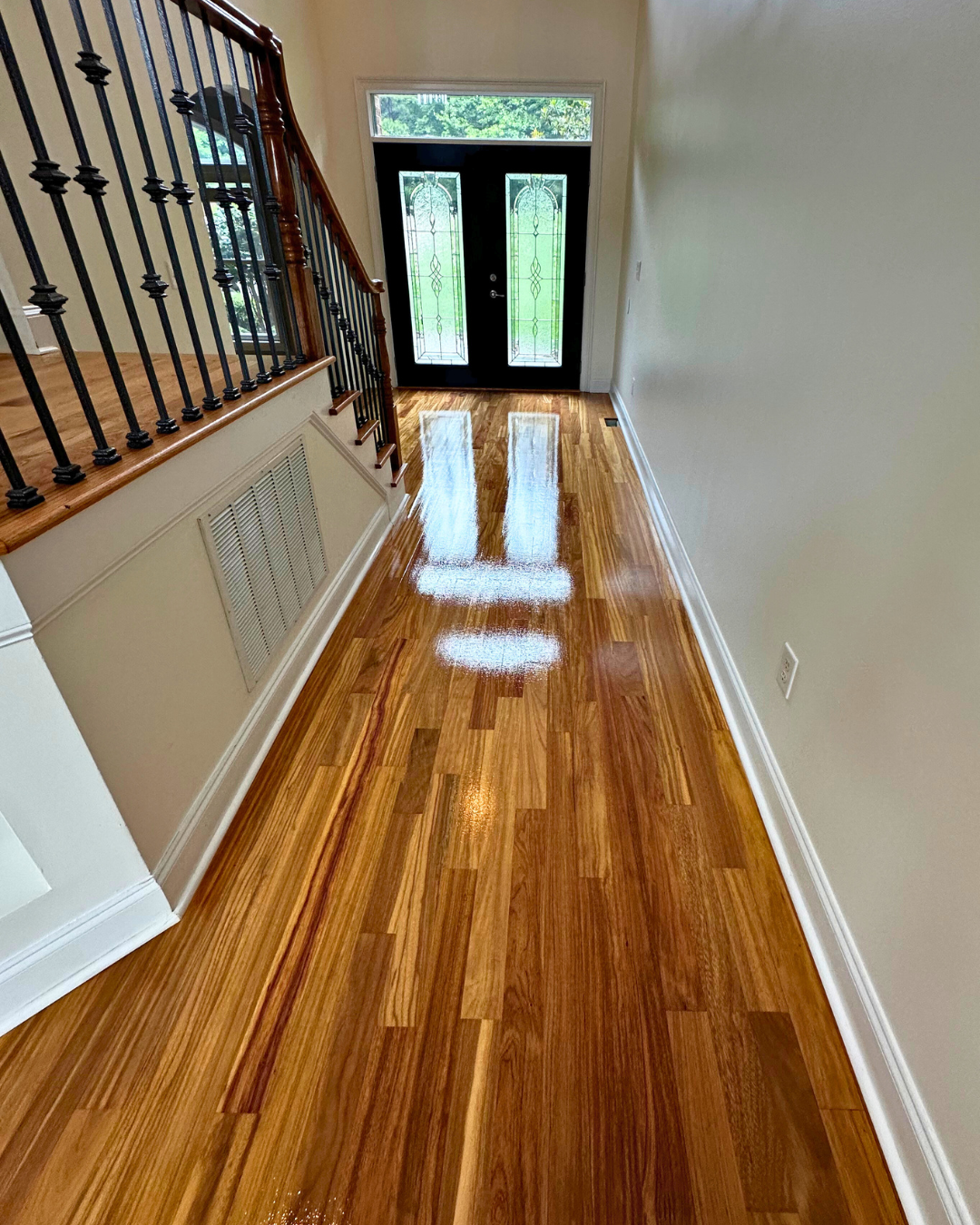 Interior view of a hallway with polished hardwood floors, a staircase with black metal balusters and a wooden handrail, and a black front door with glass panels, near a window allowing natural light.