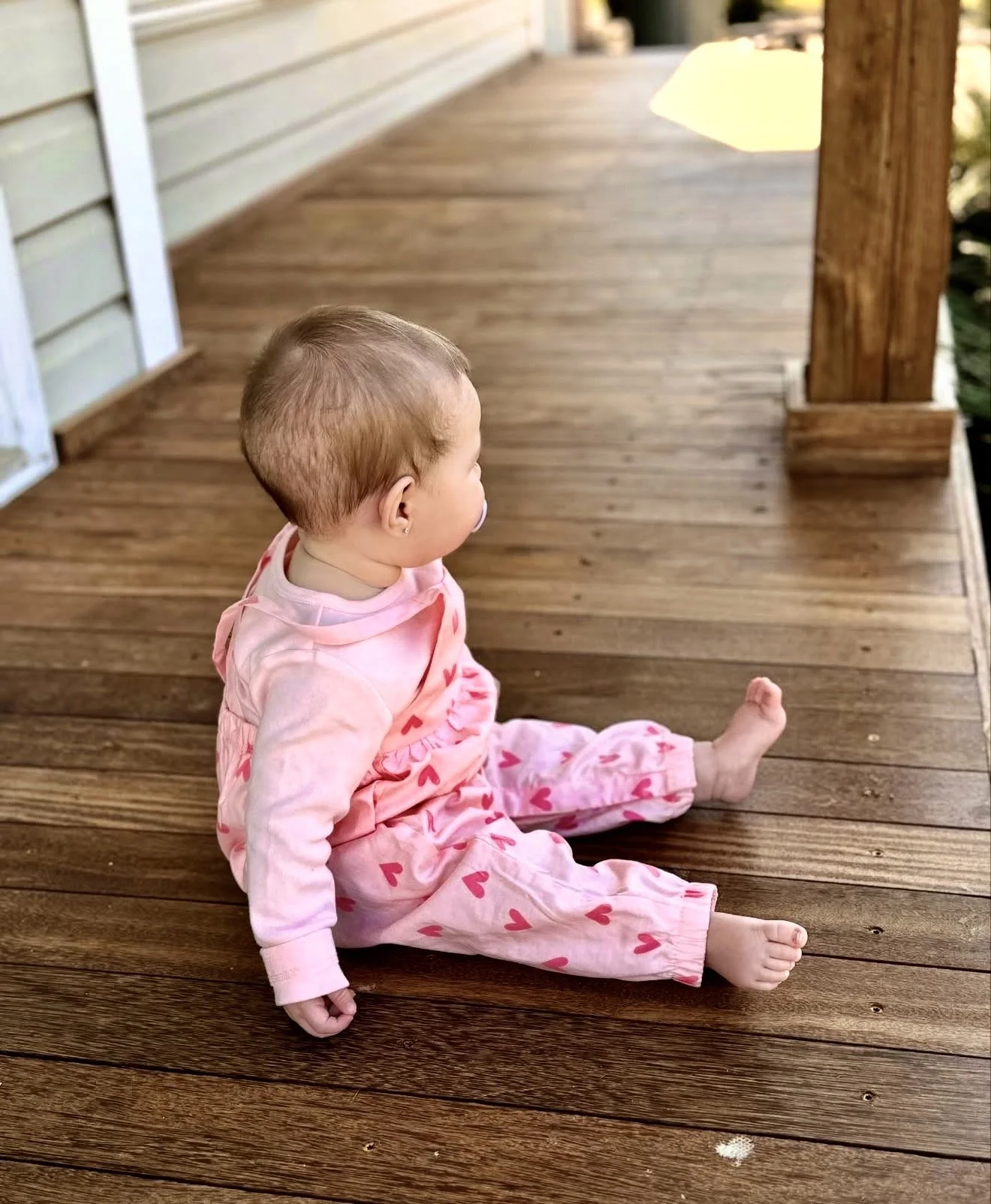 A young child with a pacifier sitting on a wooden porch, wearing pink pajamas with red hearts.