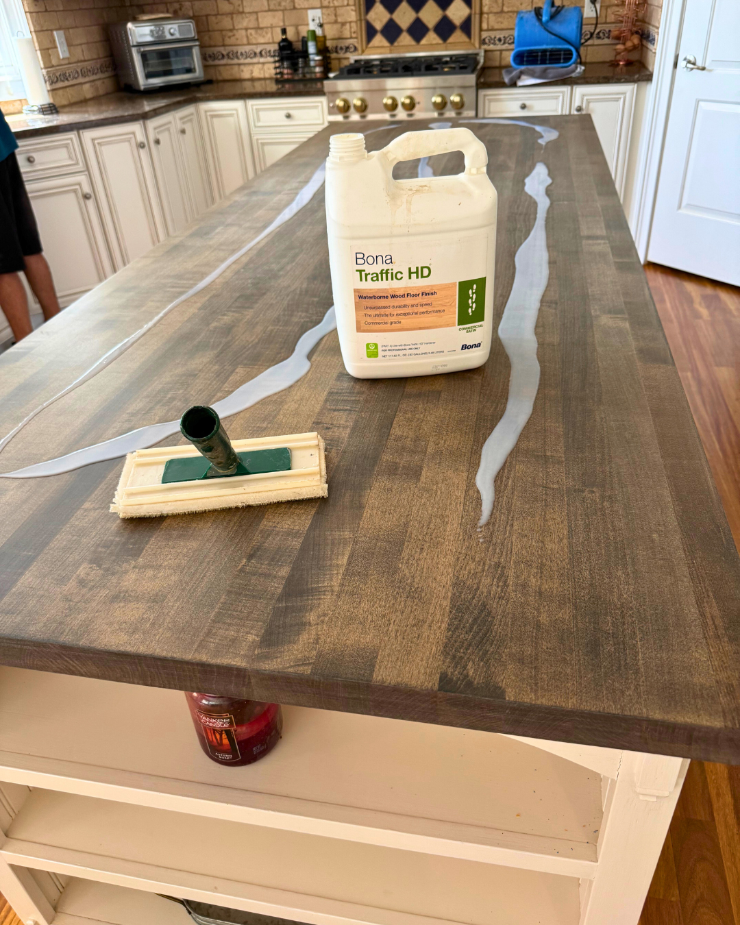A large wooden kitchen island with a bottle of Bona Traffic HD waterborne wood floor finish and a floor applicator on top, with streaks of the finish running along the surface.