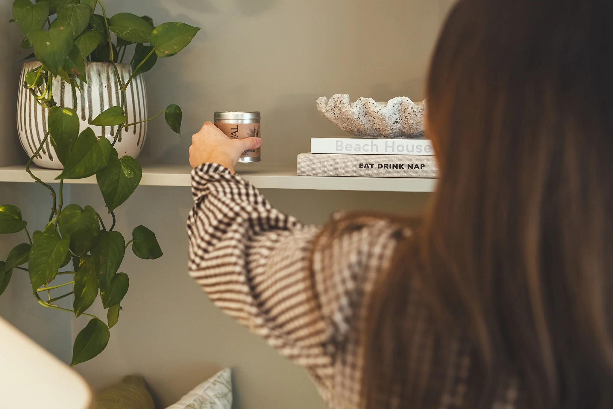 Person reaching for a candle on a white shelf next to a potted plant with green leaves. The shelf holds decorative items including a book titled 'Beach House' and another with the words 'Eat Drink Nap'.