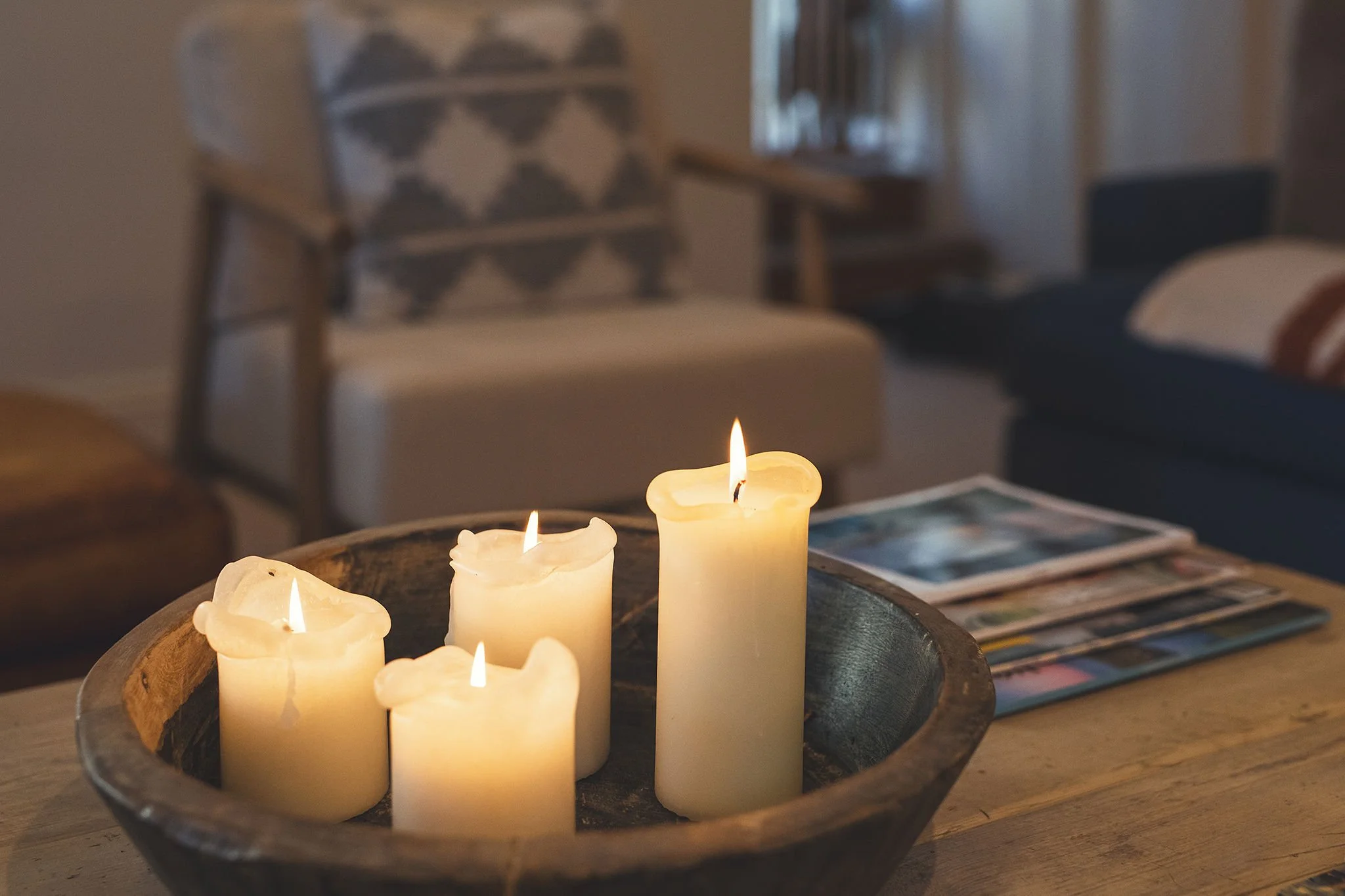 A wooden bowl holding five lit white candles on a wooden table in a cozy living room.