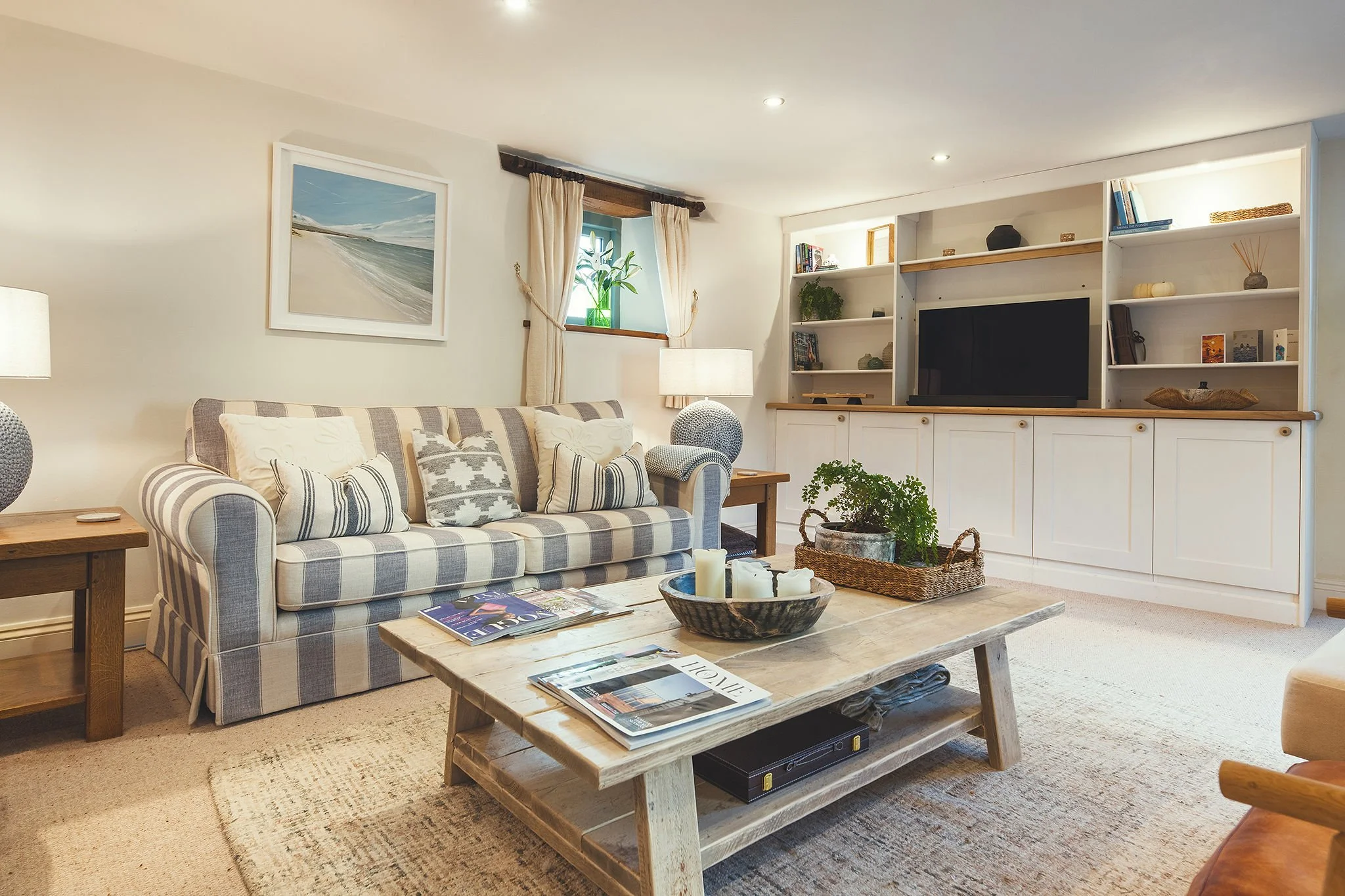 A cozy living room with a striped sofa, a wooden coffee table with magazines and candles, a side table with a lamp, a white built-in entertainment center with books and decorations, a small window with beige curtains, and wall art depicting a beach s