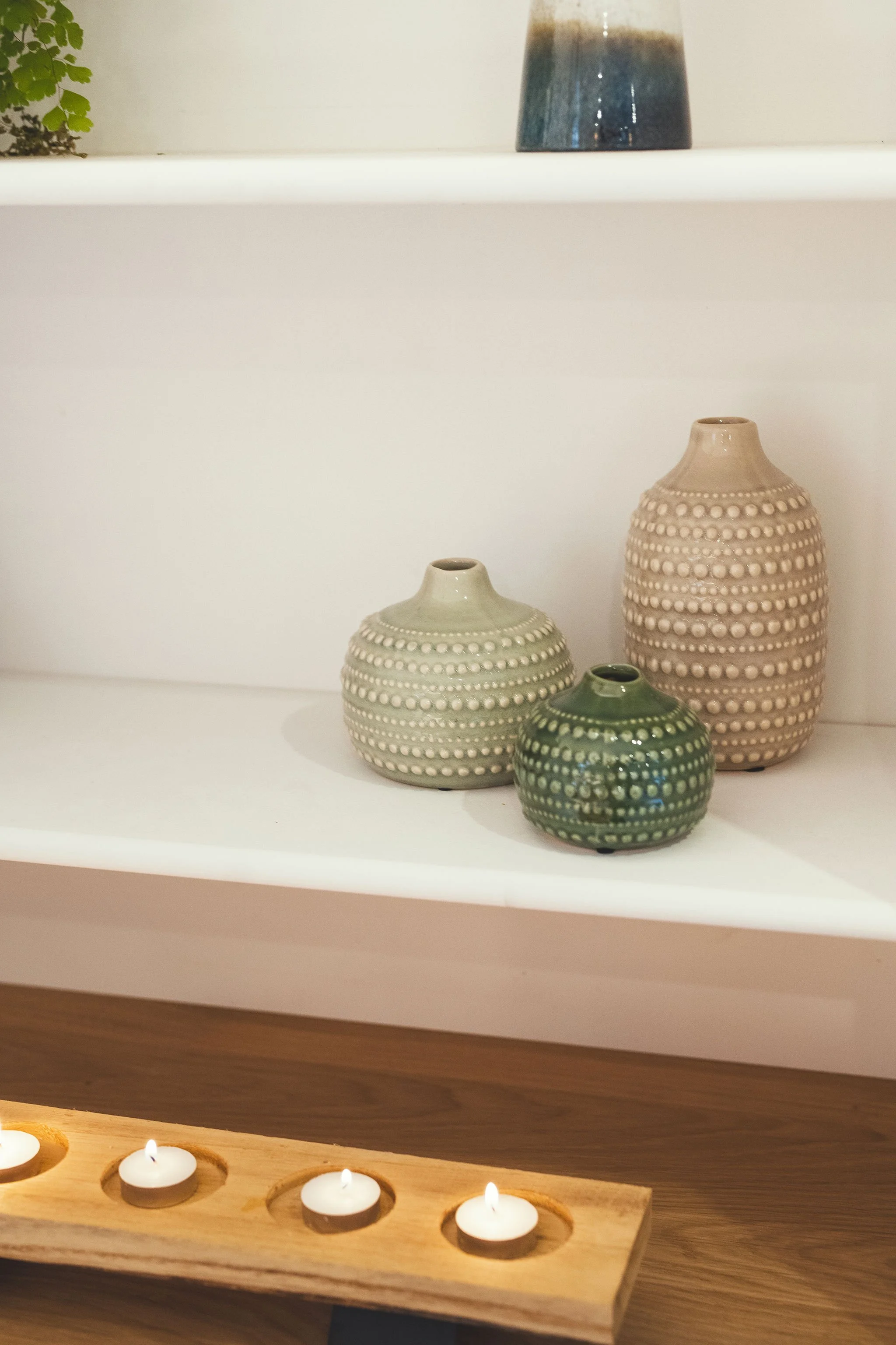 Three decorative ceramic vases of different sizes and colors on a white shelf, with a candle holder with four candles in the foreground.