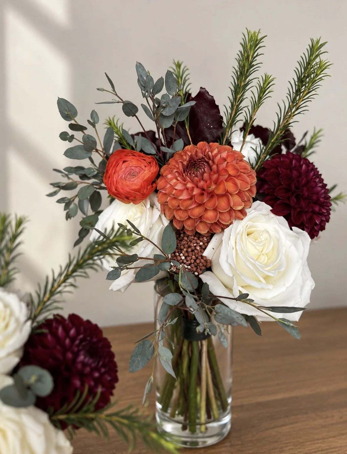A bouquet of flowers in a glass vase, featuring white roses, orange and dark red dahlias, eucalyptus leaves, and sprigs of pine.
