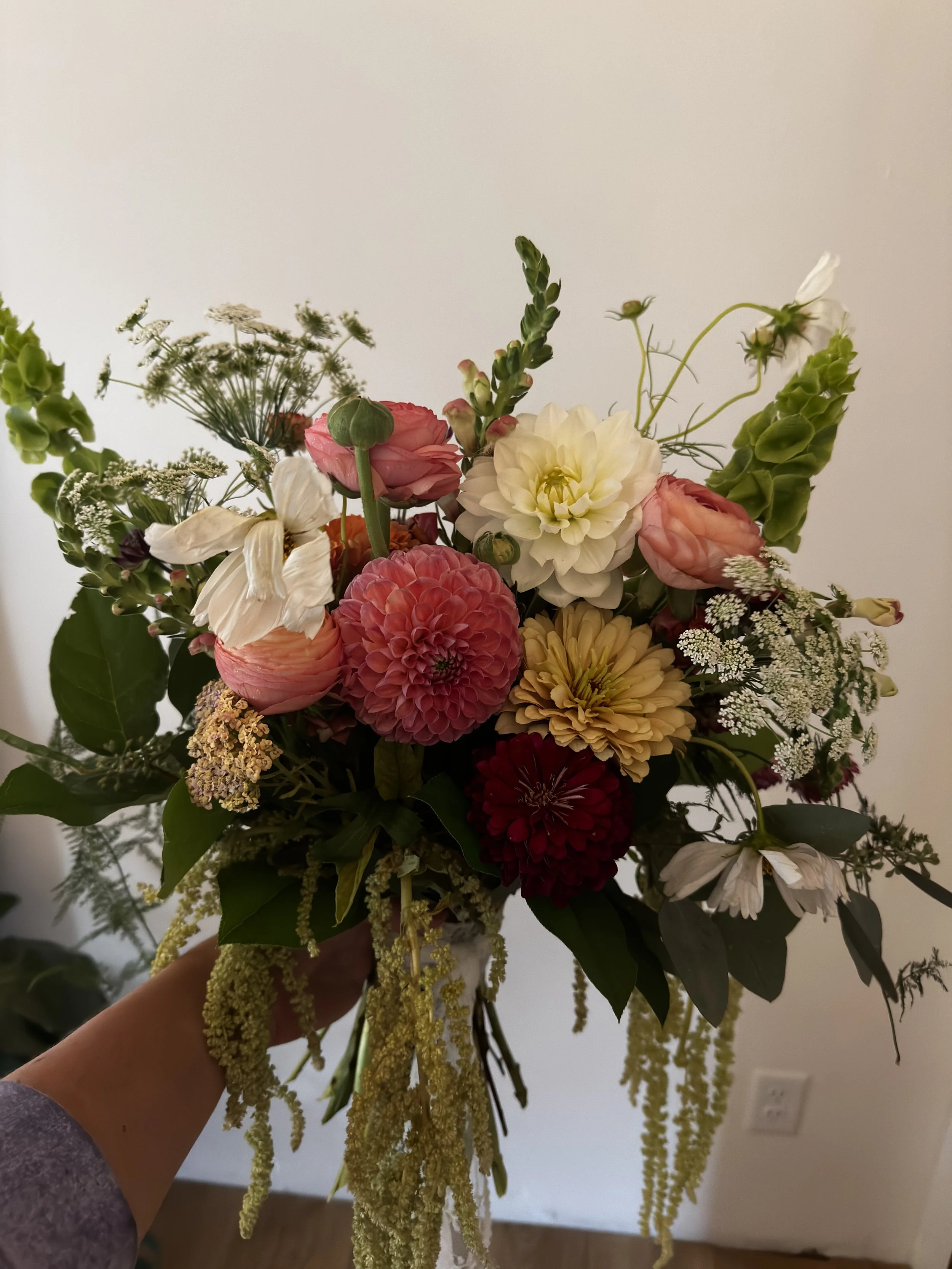 Person holding a bouquet of mixed flowers including roses, dahlias, and other greenery against a plain wall.