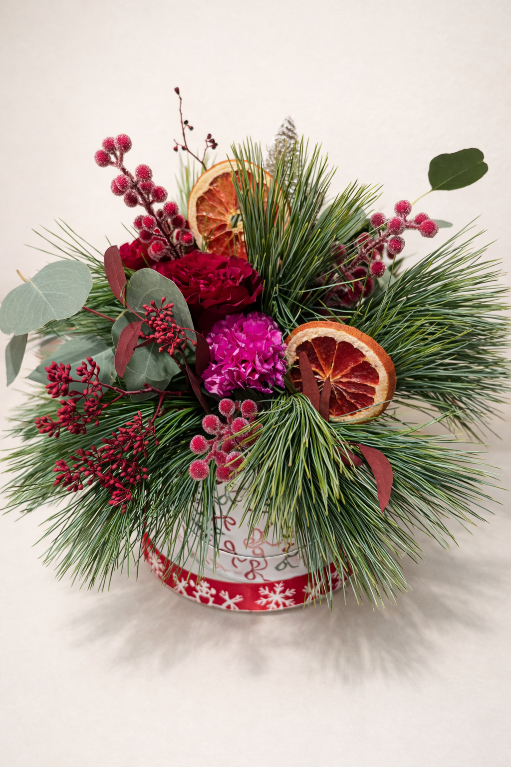 Holiday floral arrangement with pine branches, pink and red flowers, dried citrus slices, and red berries in a festive container.