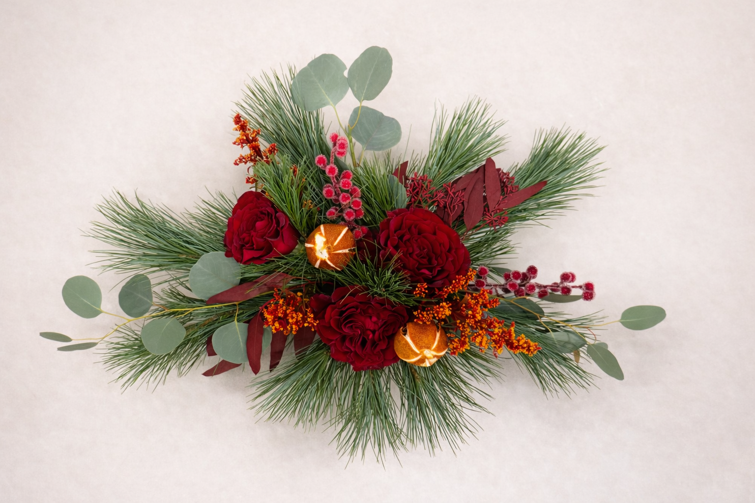 Holiday floral arrangement with red carnations, greenery, and dried orange slices on a light background.