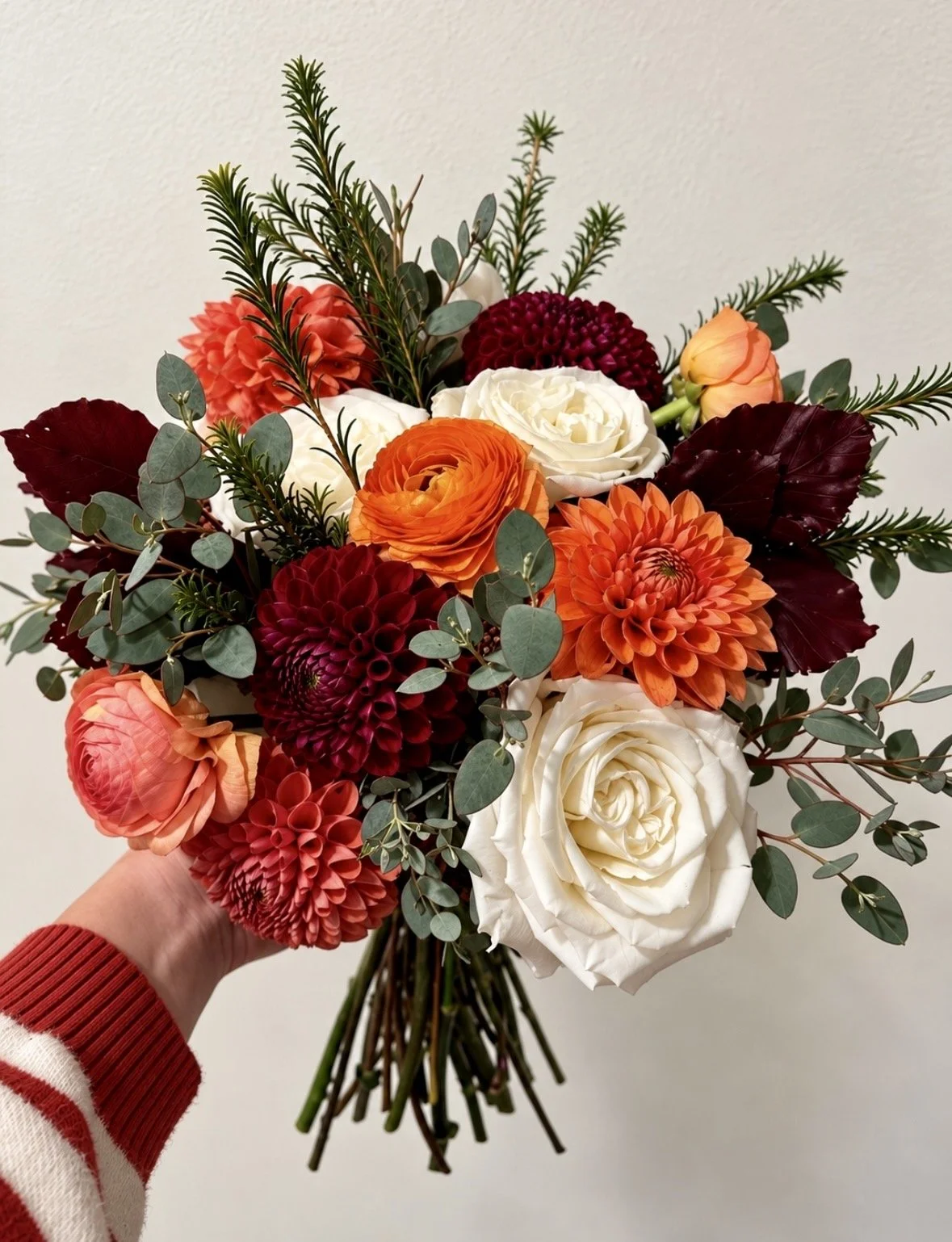 Hand holding a bouquet of colorful flowers including roses, dahlias, ranunculus, and greenery against a plain background.