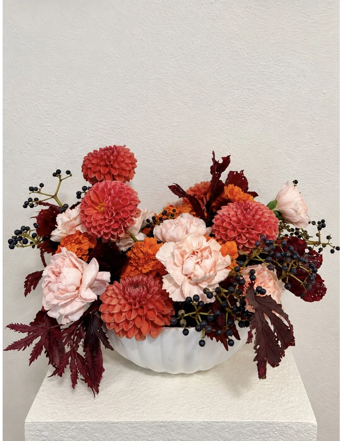 A floral arrangement in a white bowl featuring pink and orange dahlias, light pink roses, dark red leaves, and black berry stems on a white pedestal against a plain wall.