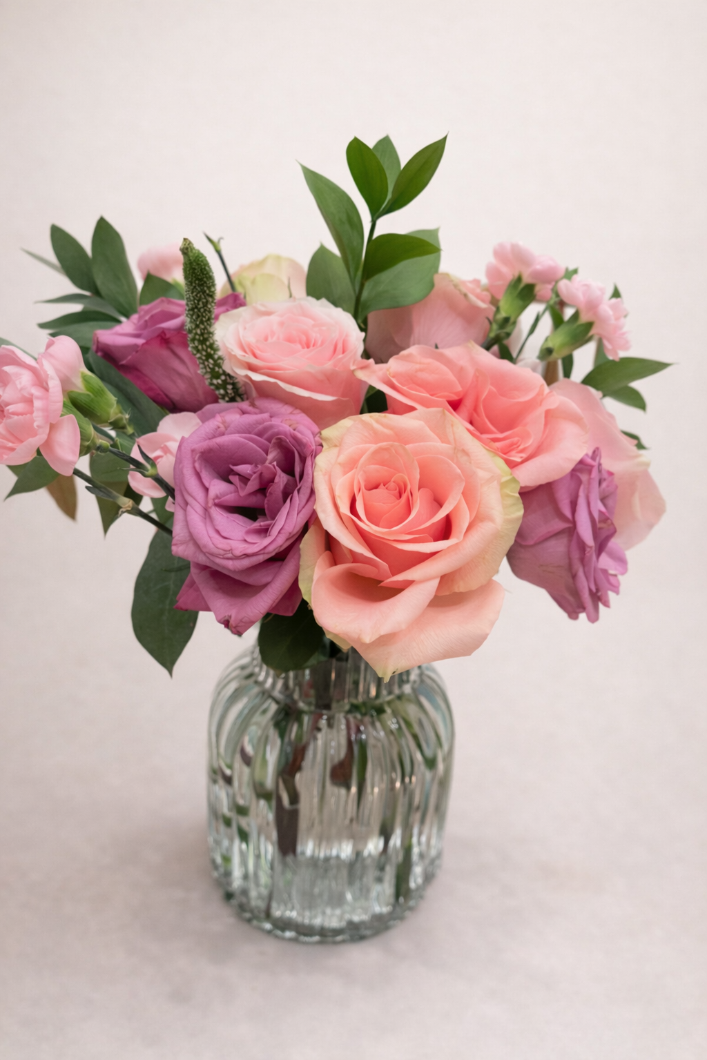 A bouquet of pink, purple, and peach roses with green leaves in a fluted clear glass vase.