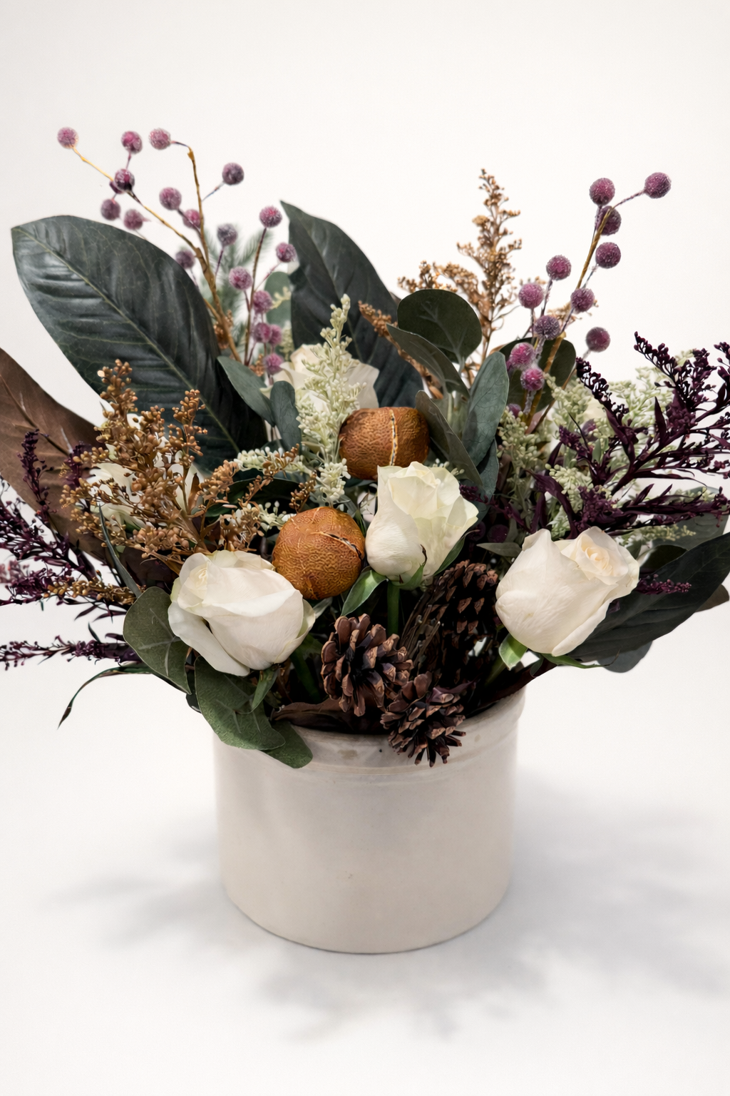 A floral arrangement in a white pot consisting of white roses, green leaves, pinecones, dried purple flowers, and two brown seed pods.