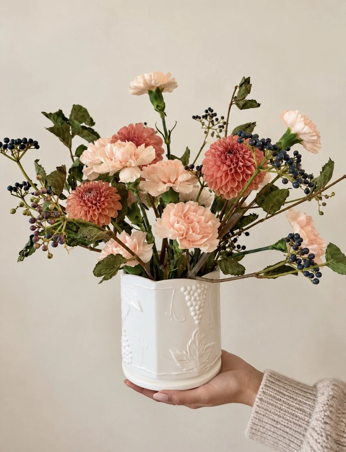 A bouquet of pink and coral dahlias, cream carnations, and sprigs of dark purple berries in a white ceramic vase with embossed grapevine design, held by a person wearing a beige knitted sweater against a plain light-colored wall.