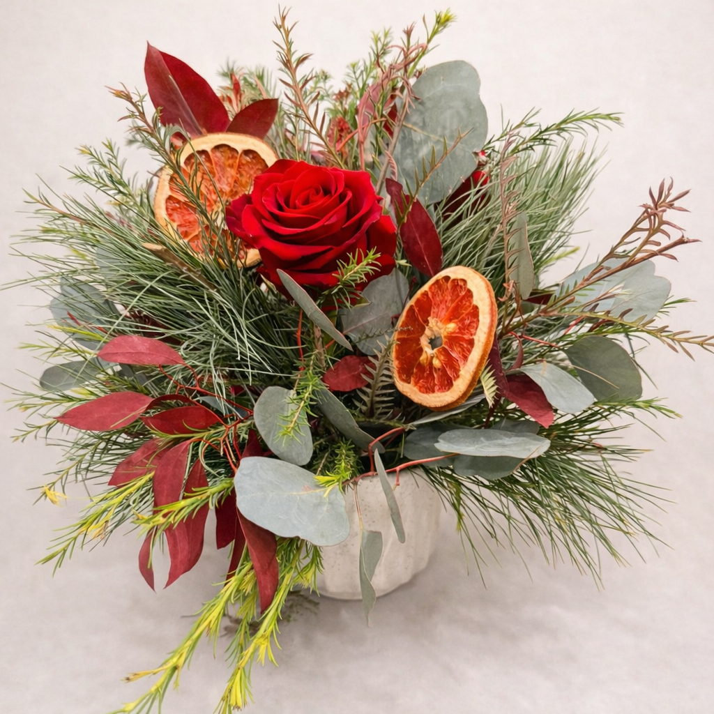 A floral arrangement featuring a red rose, dried orange slices, and various green leaves and foliage in a white vase.