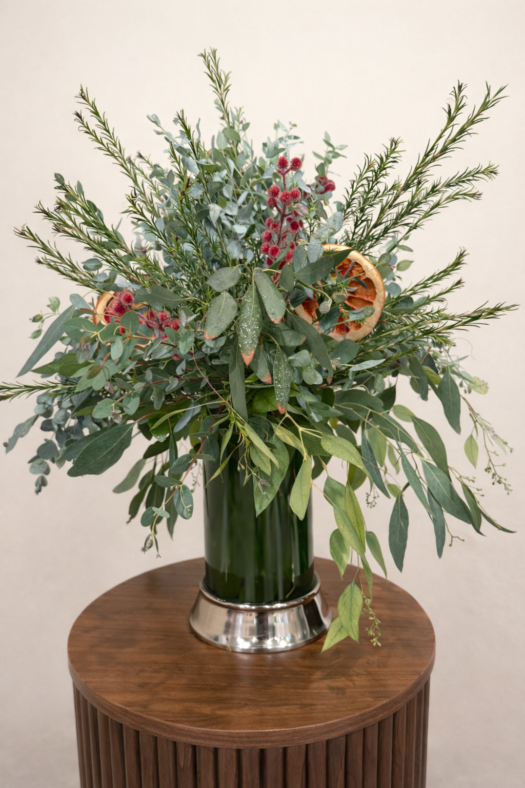 A bouquet of green foliage and red berries in a clear vase on a wooden table.