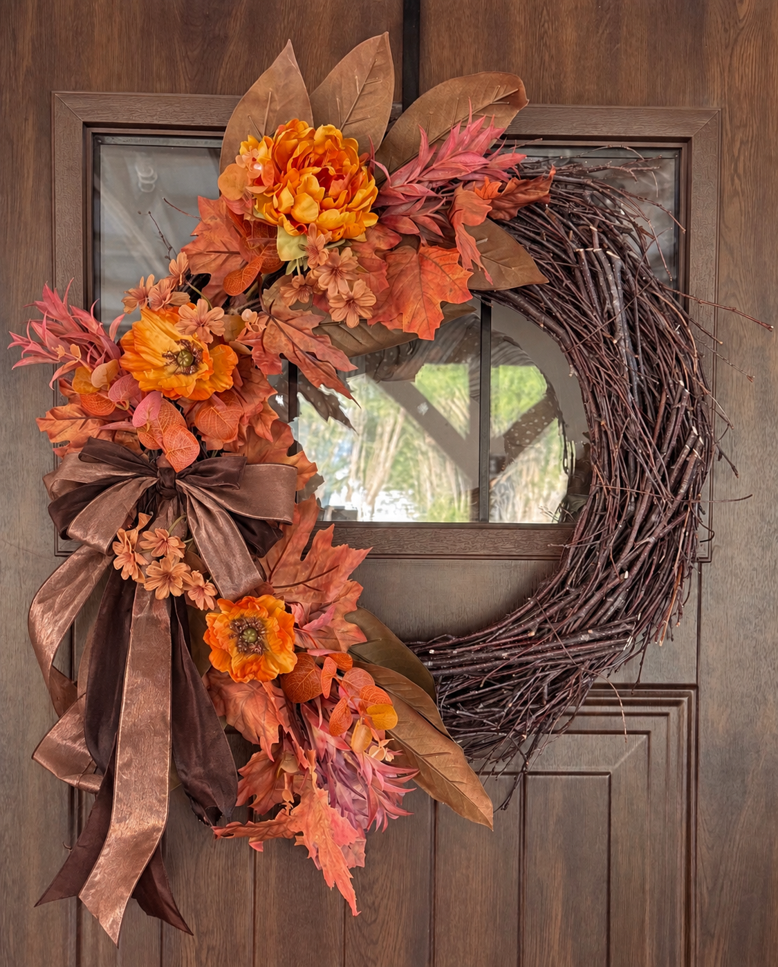 Fall-themed wreath made of intertwined branches, decorated with orange and yellow flowers, pink and orange leaves, a brown ribbon, and bows, hanging on a wooden door.