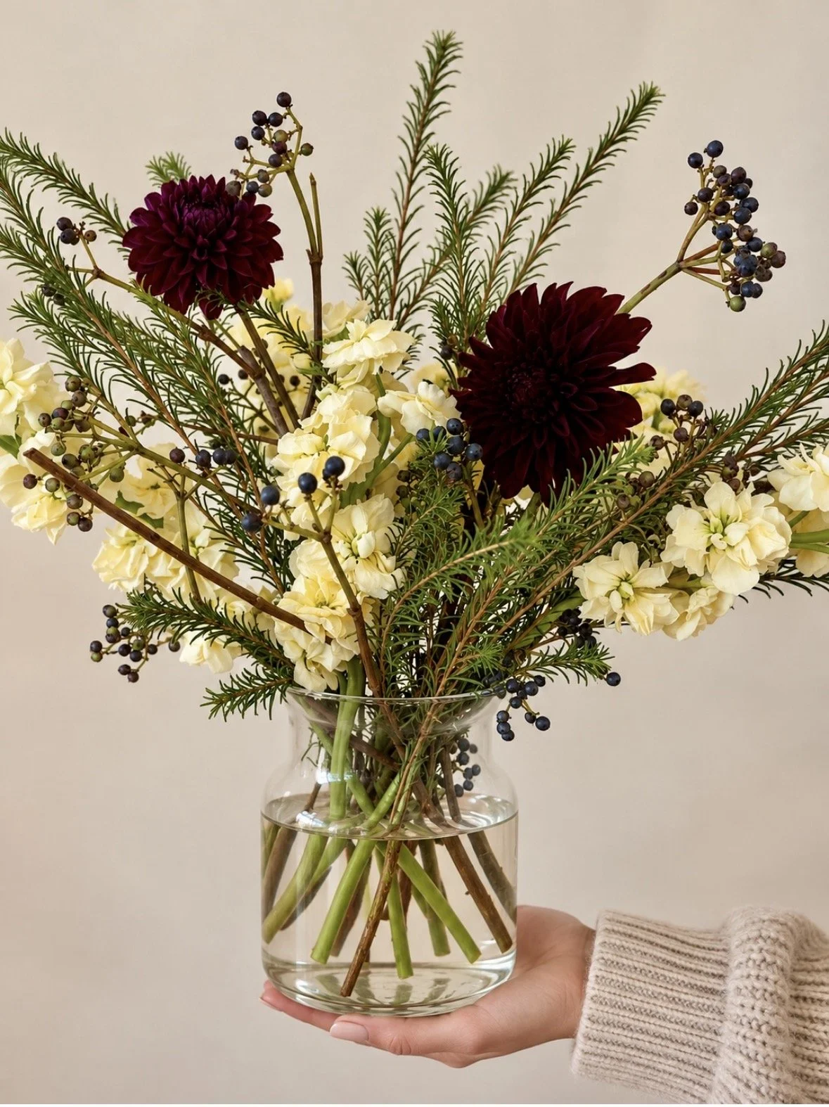 Hand holding a clear glass vase with a floral arrangement of white flowers, dark red flowers, green foliage, and small dark berries.