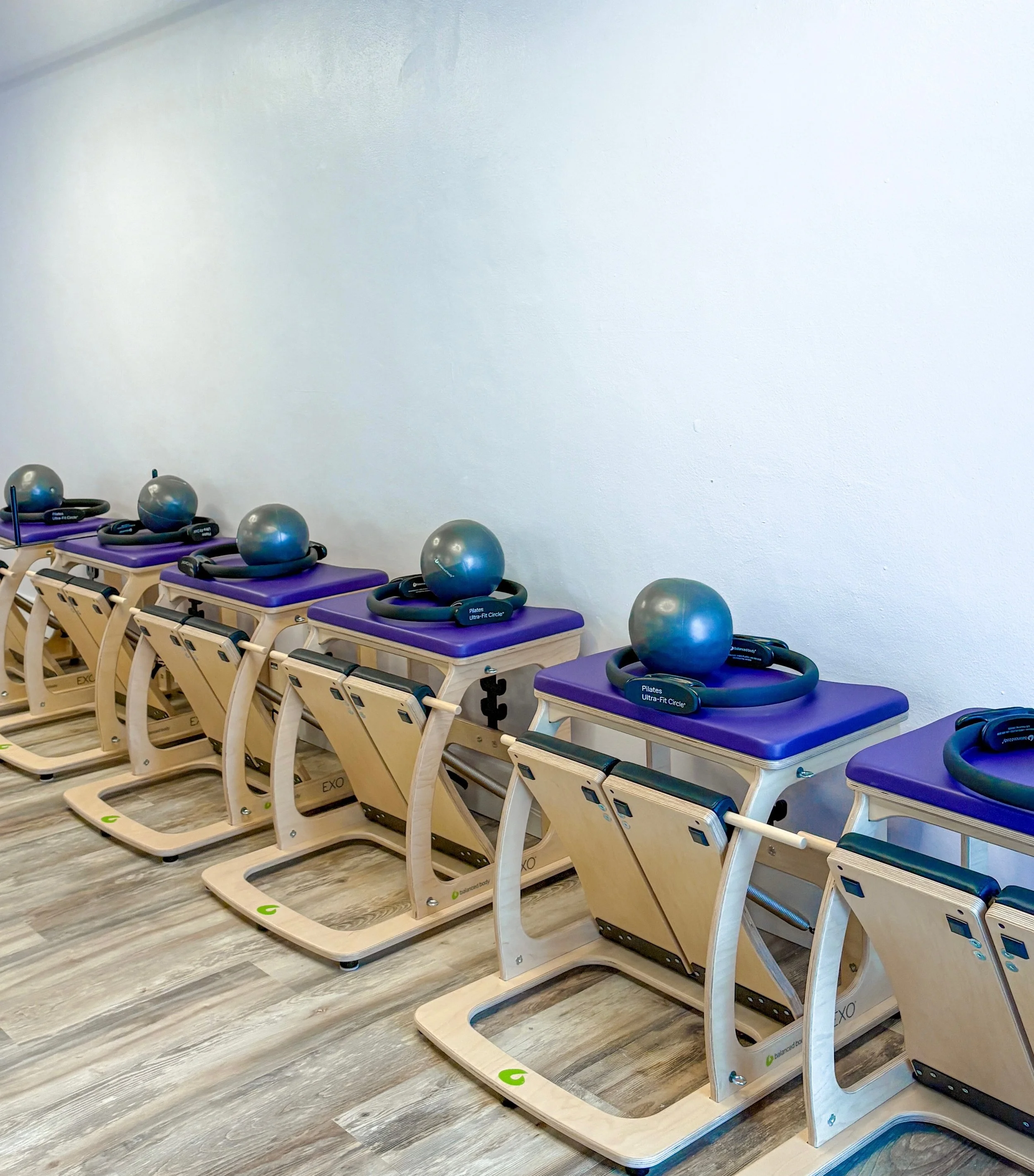 A row of Pilates reformer machines with purple cushioned surfaces and silver balls on top, set against a plain white wall in a fitness studio.