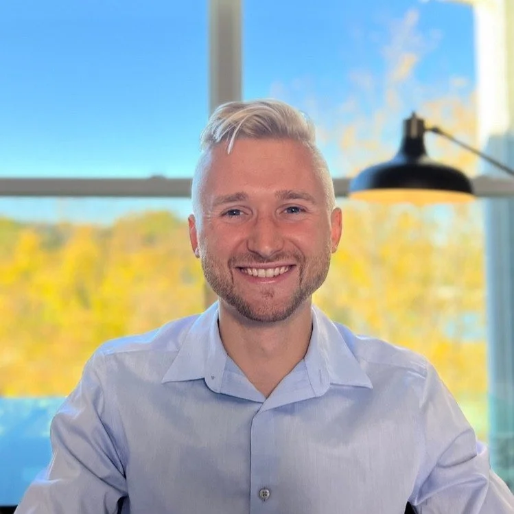 A smiling man with blonde hair and a beard, wearing a light blue button-up shirt, sitting indoors in front of a window with a view of trees with fall foliage and a blue sky.