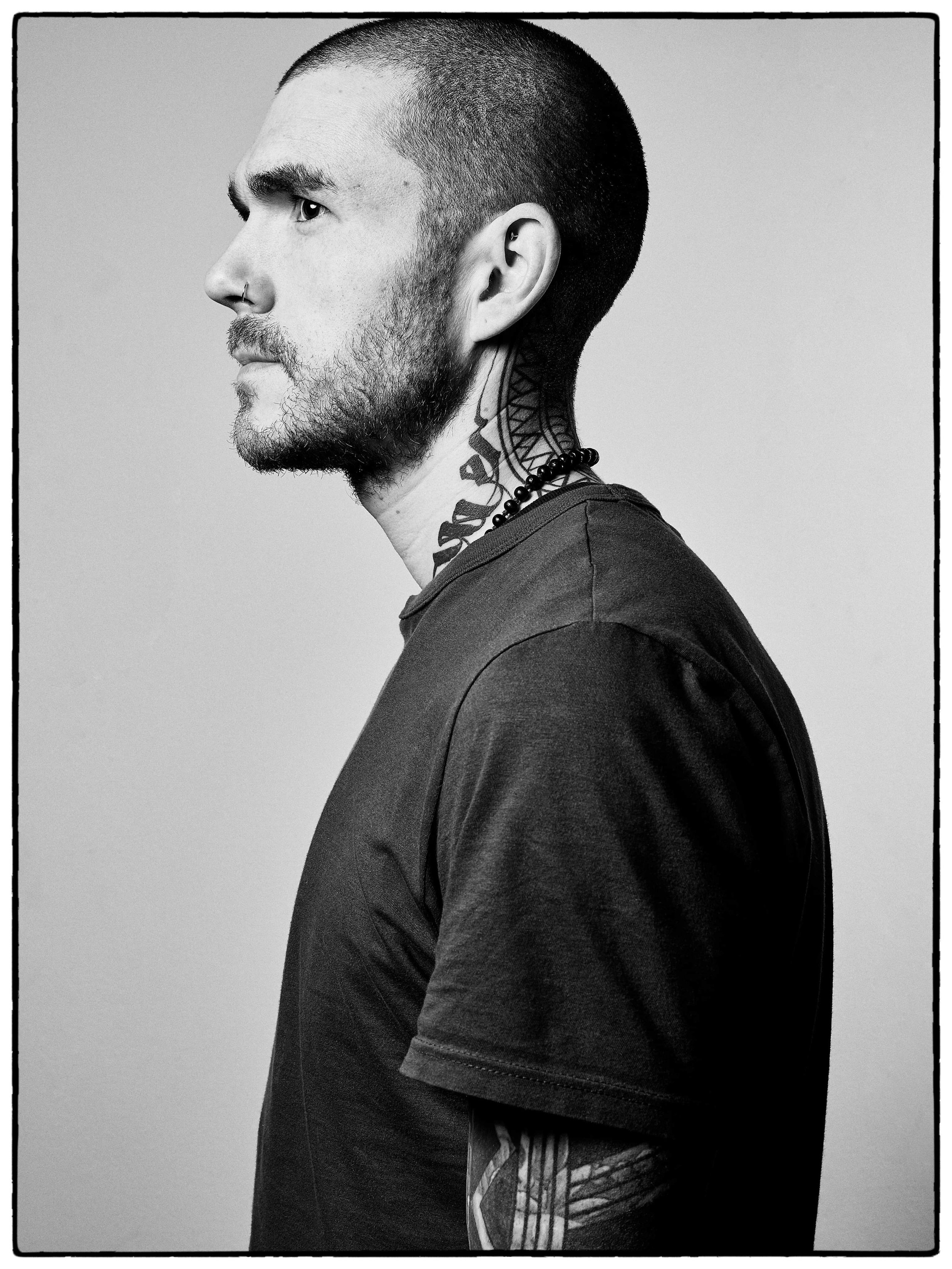 Side profile of a man with a shaved head, beard, and tattoos on the neck and arm, wearing a dark t-shirt and a beaded necklace, against a plain background.