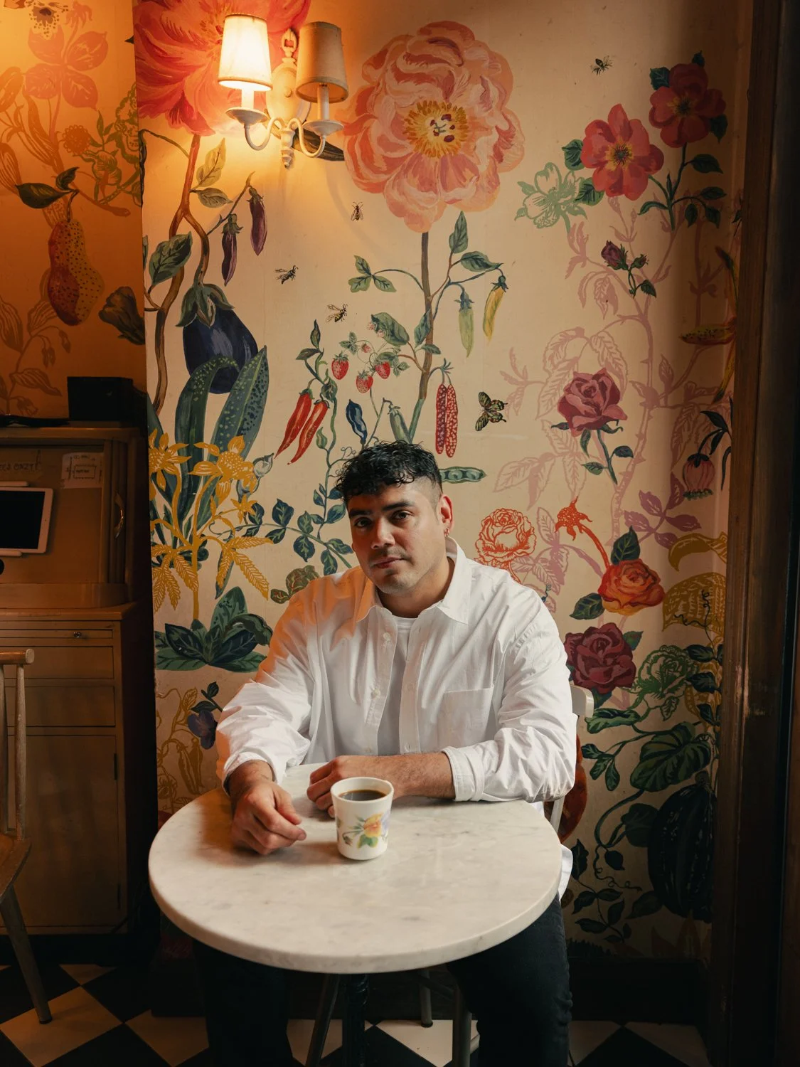 A man sitting at a small round table with a cup of coffee, in front of a colorful floral mural wall inside a cozy cafe with warm lighting.