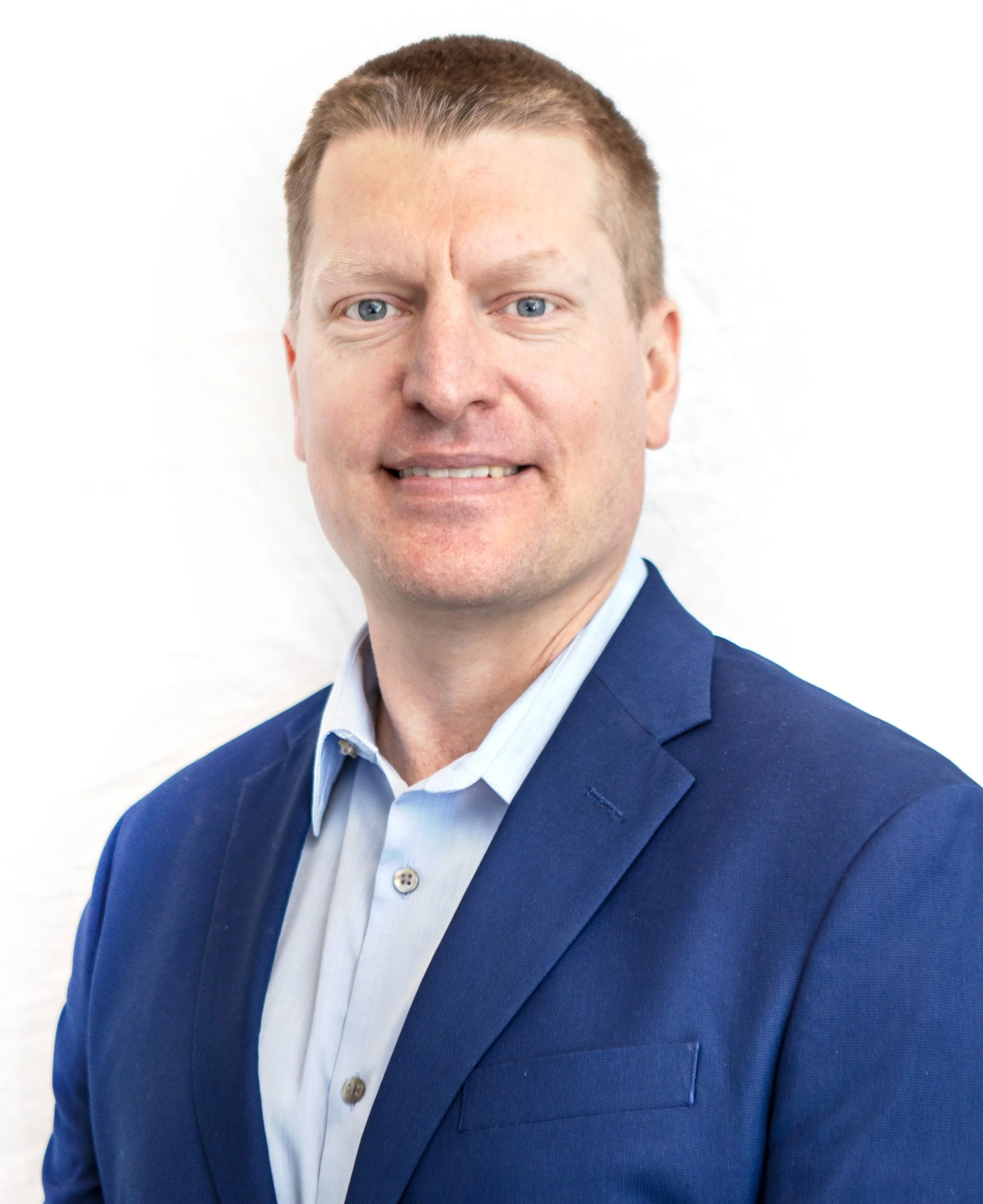 A man is smiling in front of white background, wearing a blue dress shirt under a darker blue suit jacket.