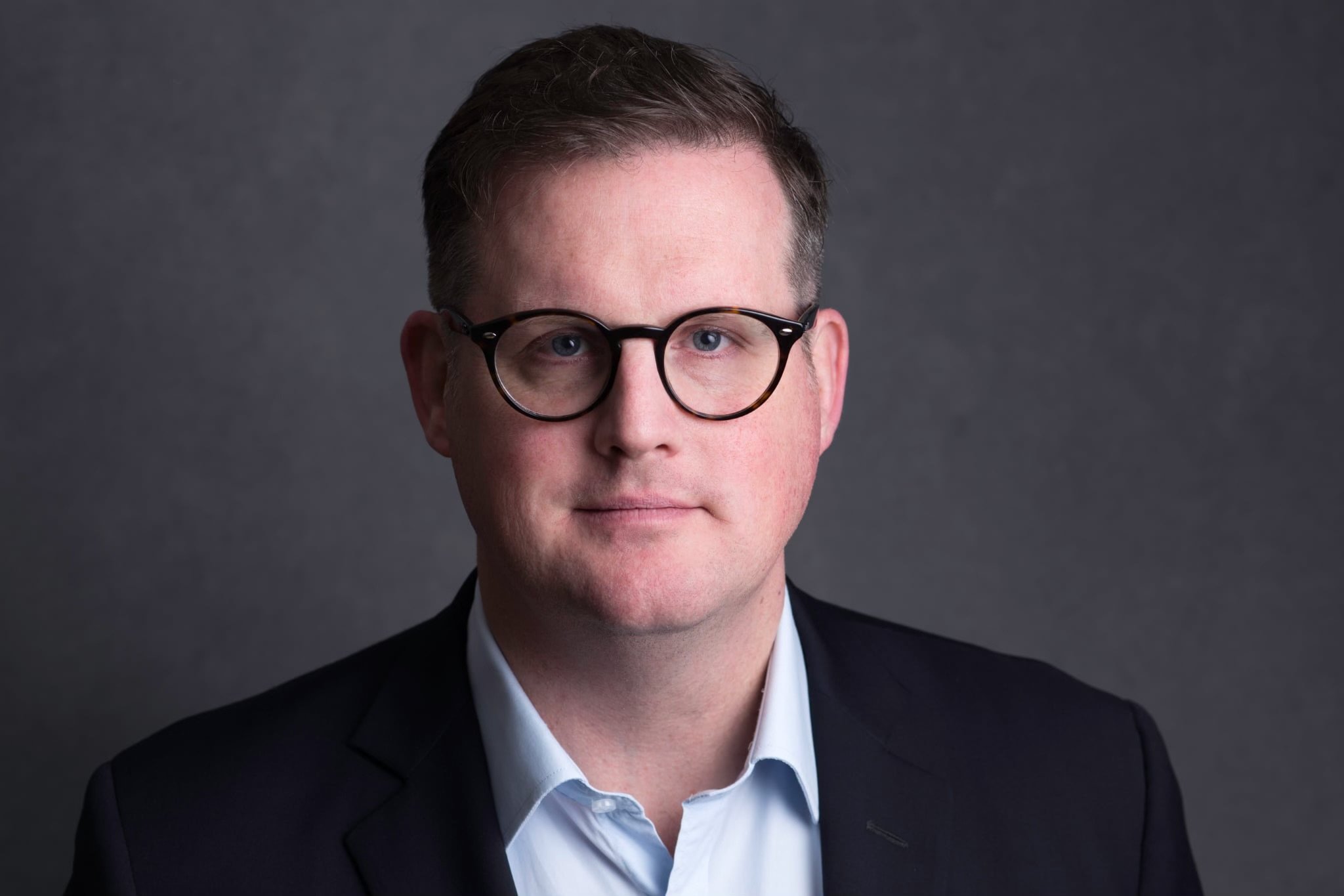A man with glasses is staring right into the camera with a neutral expression. He is wearing a blue collared shirt under a black jacket.