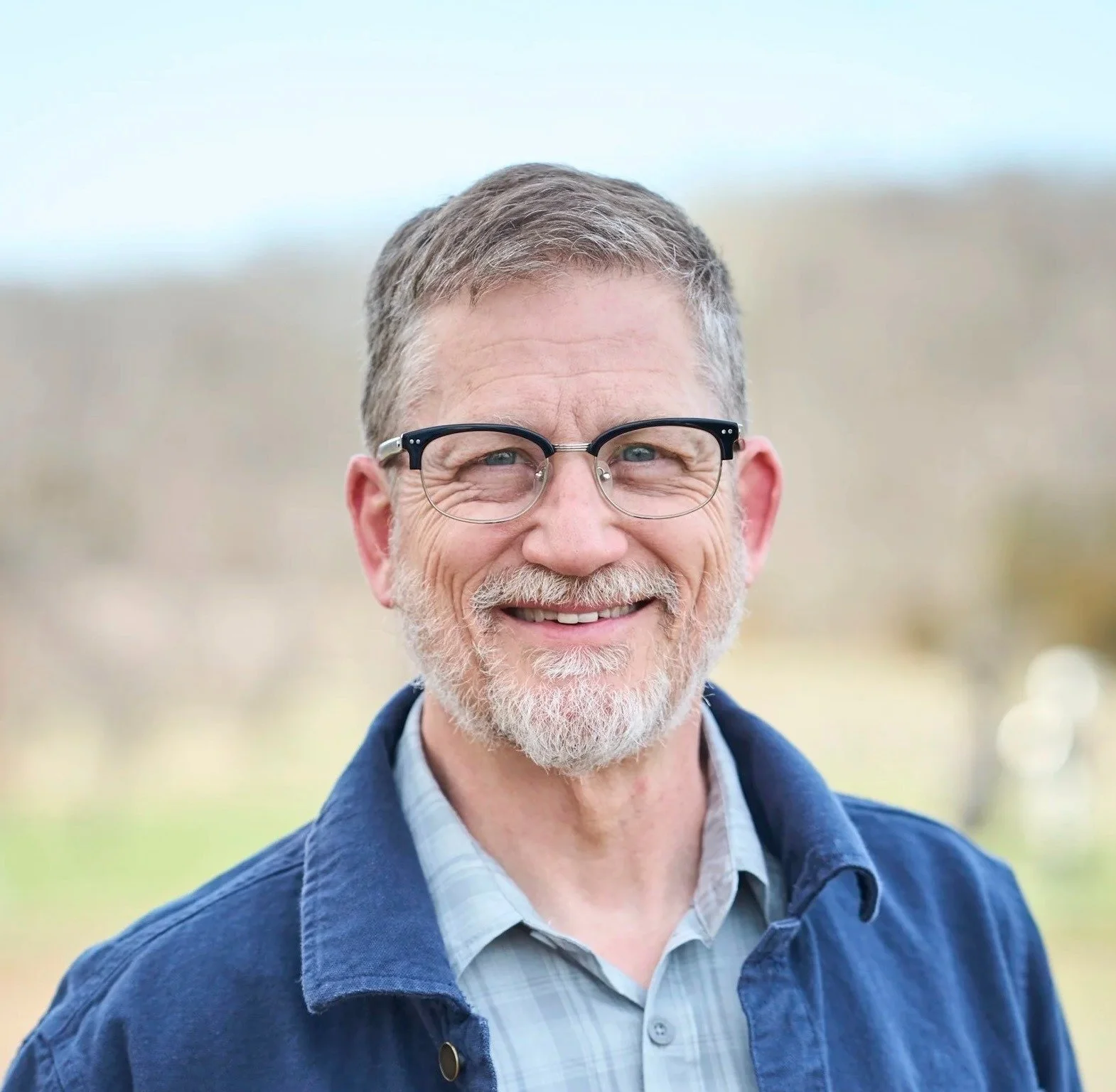 A man with glasses, a blue button up, and a blue jacket is smiling close to the camera for an outdoor headshot.