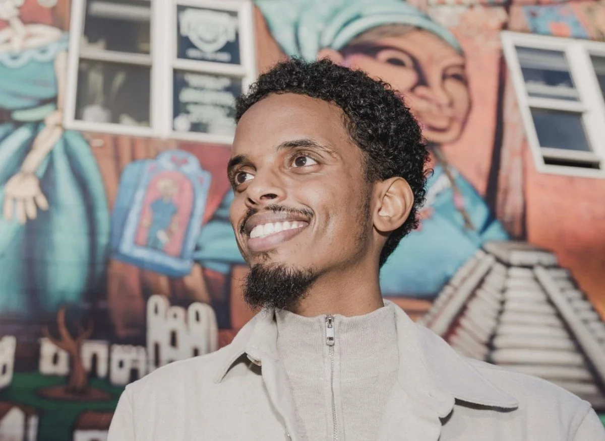 A man with curly black hair and facial hair wearing a khaki jacket is smiling off camera in front of a mural.