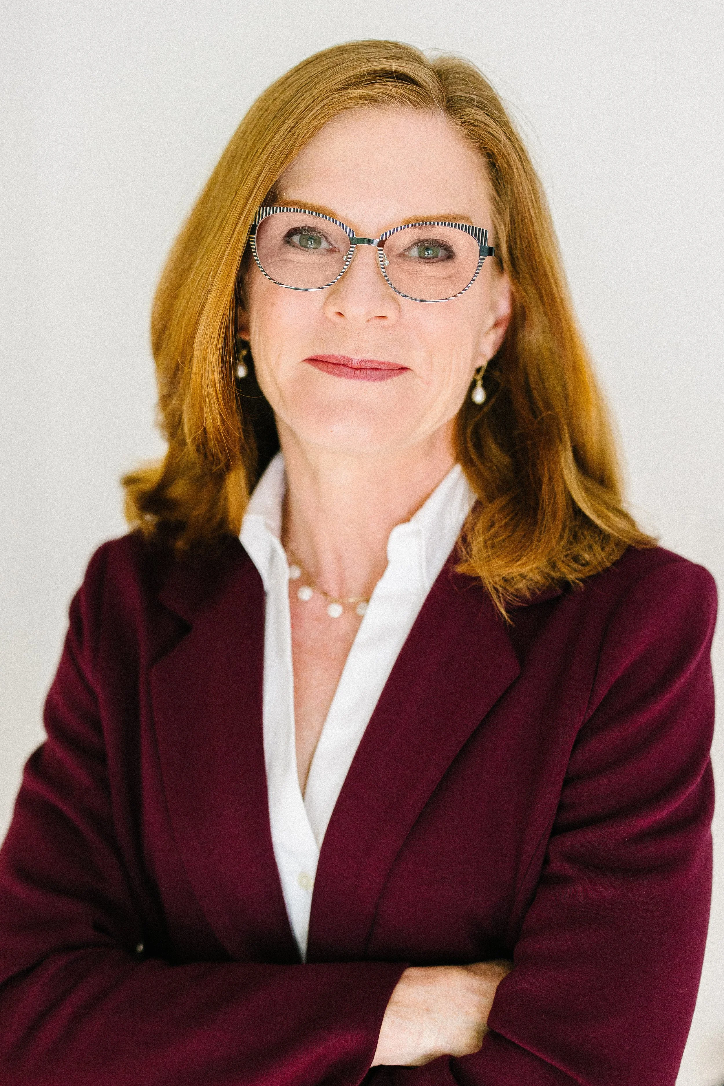 A woman with shoulder length red hair is standing with her arms crossed in a headshot. She is wearing a maroon jacket over a white button up shirt and glasses.