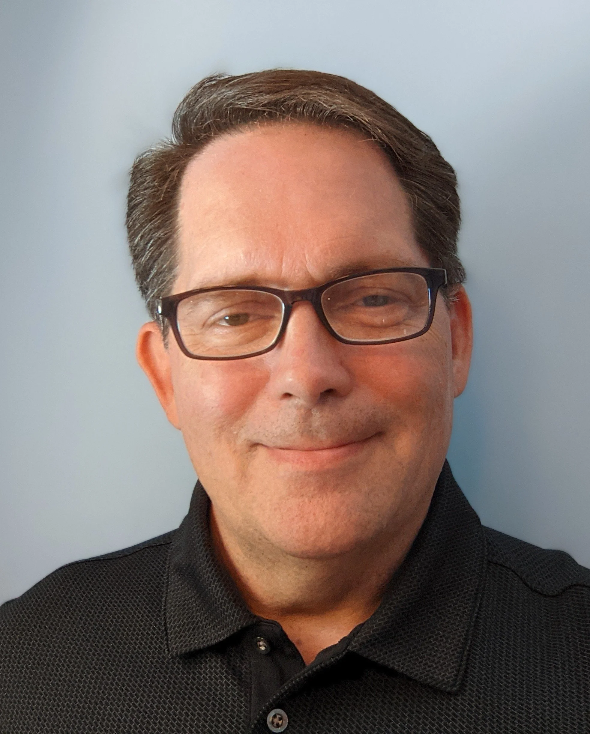 A selfie of a man in a black polo shirt has a closed lip smile at the camera. He is wearing black glasses and has brown hair.