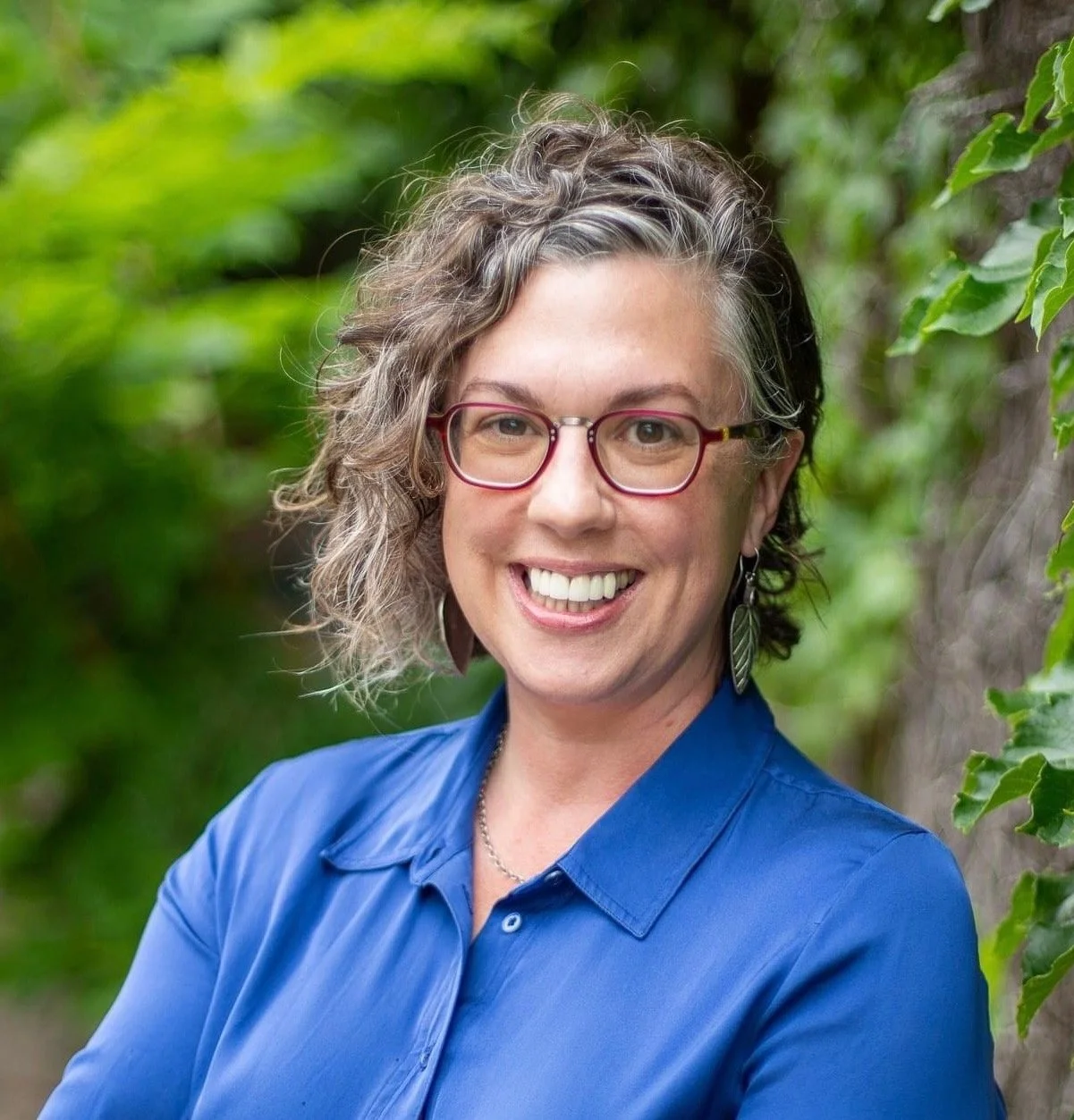A woman with curly gray hair and red glasses is wearing a blue dress shirt and leaning against a tree for a casual headshot.