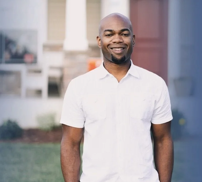 A man wearing a white collared shirt is smiling against a city backdrop.