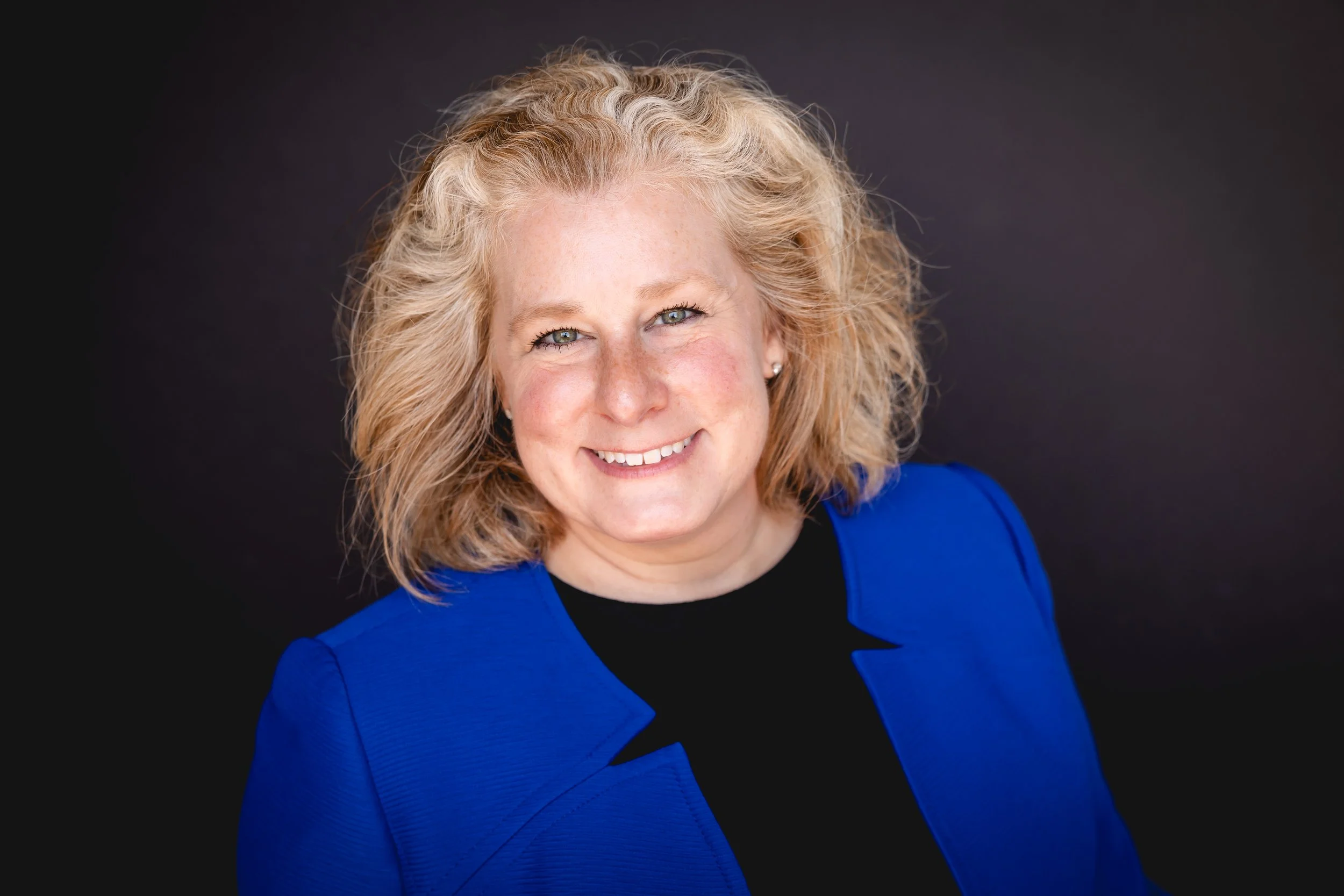 A woman with blonde shoulder-length hair is smiling into the camera in a headshot. She is wearing a blue blazer over a black shirt.