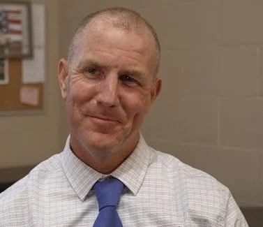 A man with dimples is wearing a blue tie and white dress shirt is smiling slightly off camera.