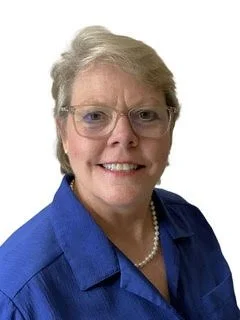 A woman with blond hair, glasses, a pearl necklace, and a blue suit is in a headshot with a white background.