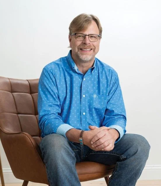 A smiling man in glasses and a blue dress shirt is seated in a brown leather chair with his hands folded in his lap.