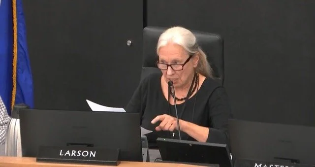 A woman with long white hair tied back in a ponytail and is wearing all black is seating at a desk that holds a name plate that reads "Larson". She is addressing an audience, as she is speaking into a microphone and pointing with one hand.