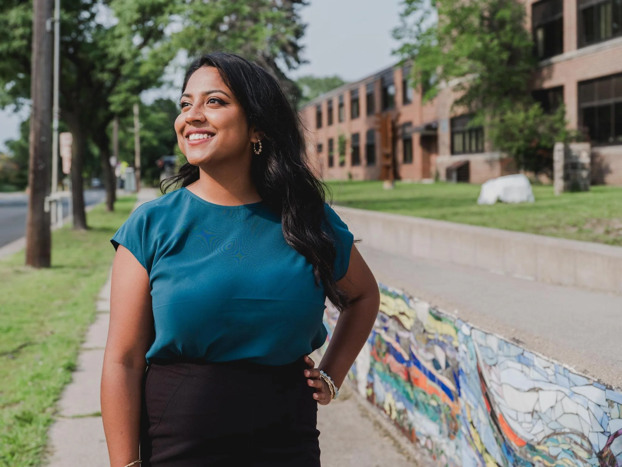 A woman with long black hair, a blue shirt, and black skirt is smiling off camera. She is in front of a city street with buildings, trees, and grass.