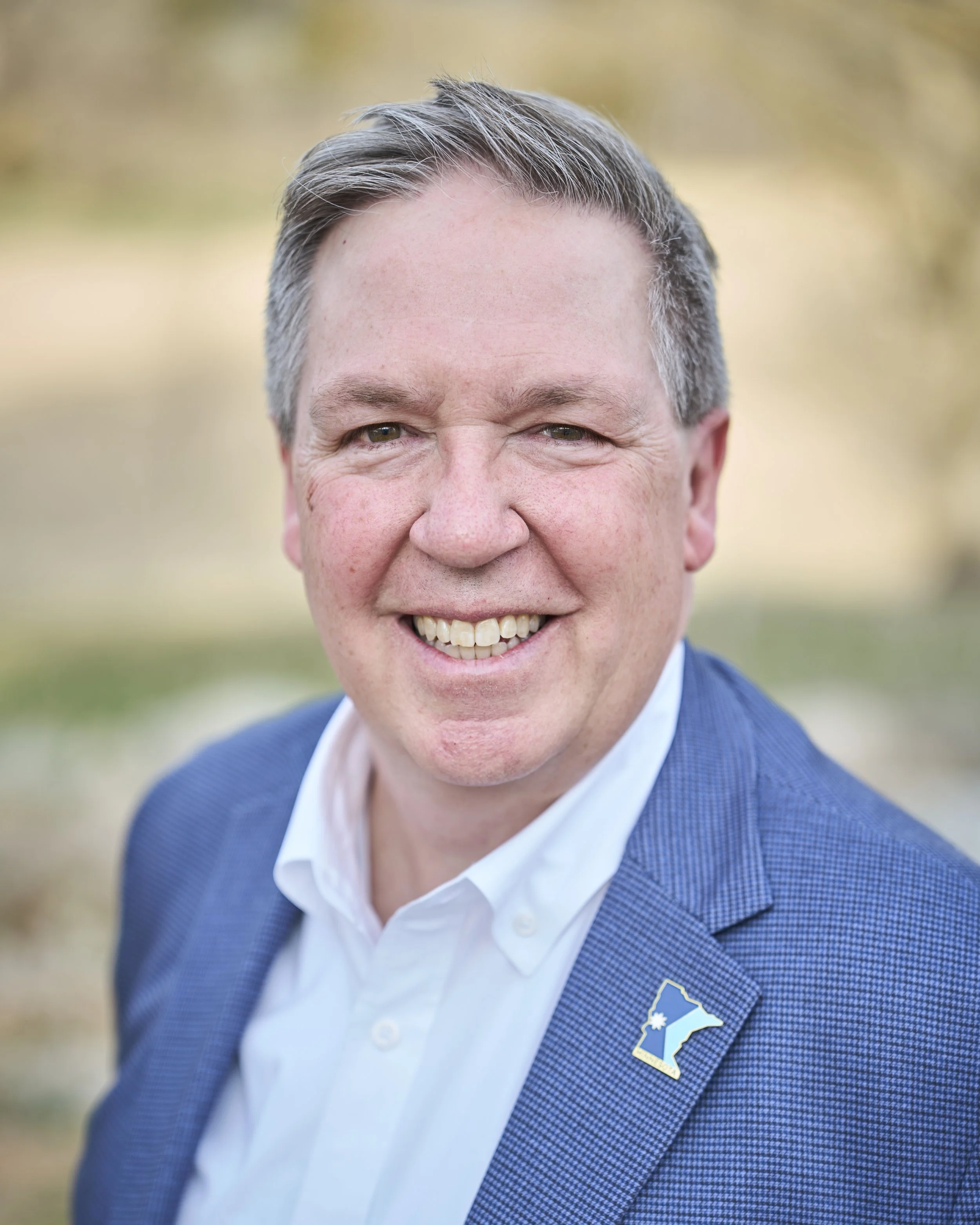 A smiling man with short gray hair and a blue suit is smiling outdoors close to the camera for a headshot.