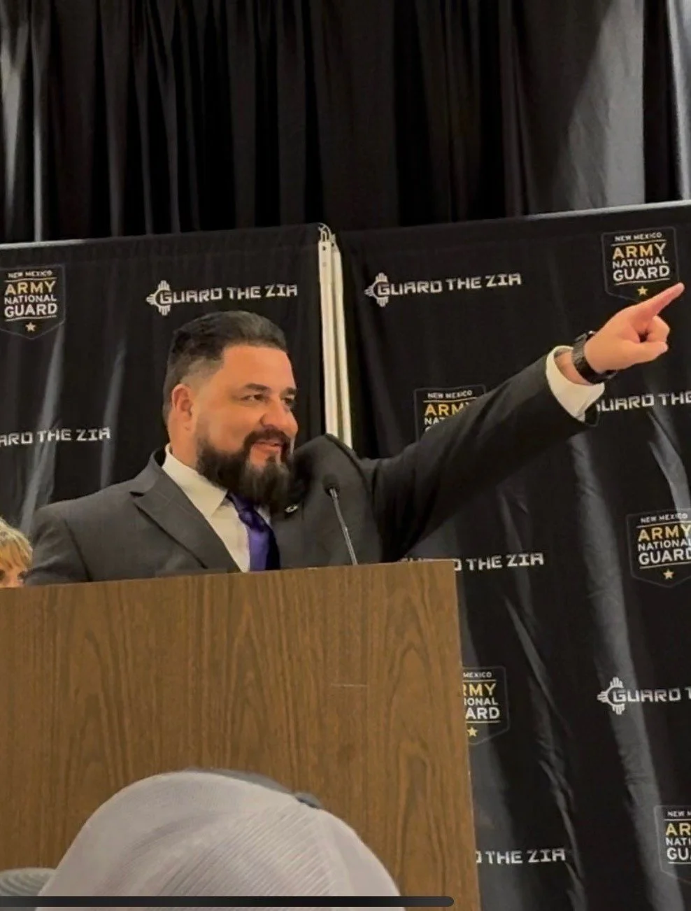 A man at a podium pointing to a banner that says 'Guard the Zia' with the New Mexico Army National Guard logo, also standing in front of a black curtain.