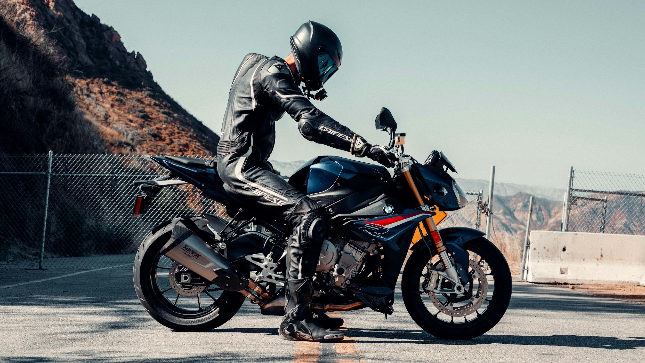 Motorcycle rider in black riding gear and helmet preparing to ride on an empty road with mountains in the background.
