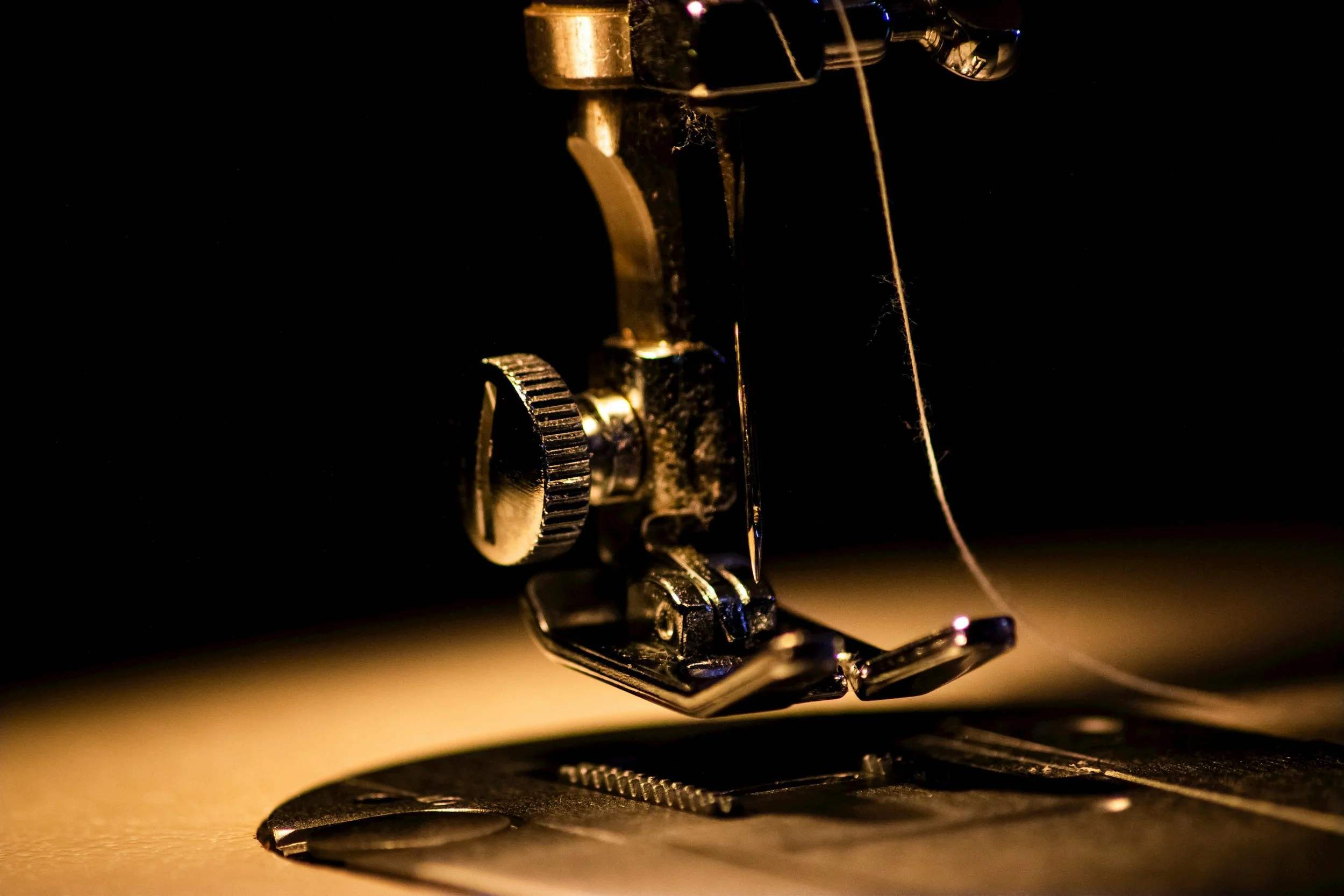 Close-up of a sewing machine needle and presser foot, with a thread passing through, set against a dark background.