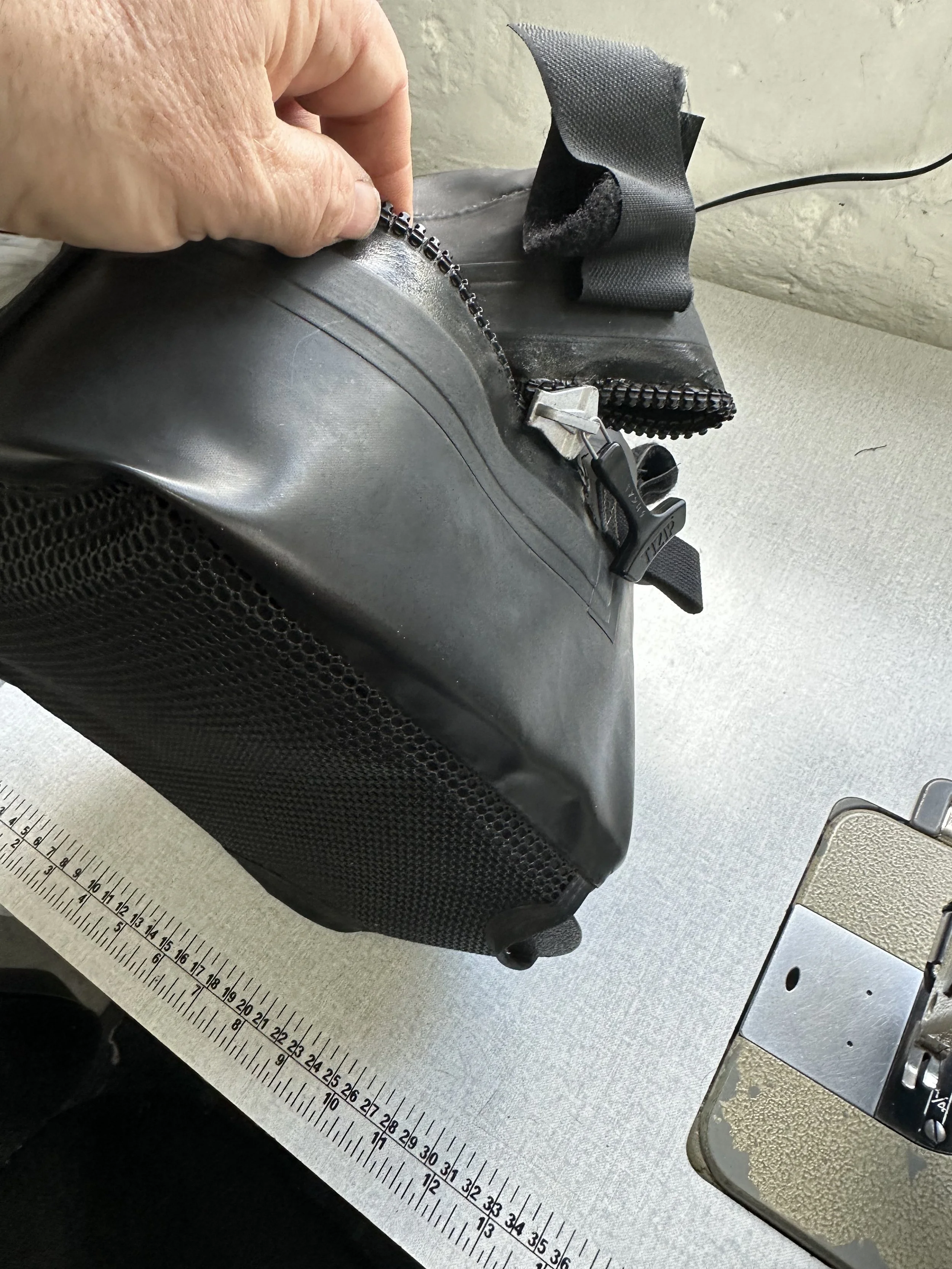A close-up of a black, textured, hard-shell bag with a zipper being opened by a person's hand. The bag is placed on a surface with a ruler and a metal device nearby.