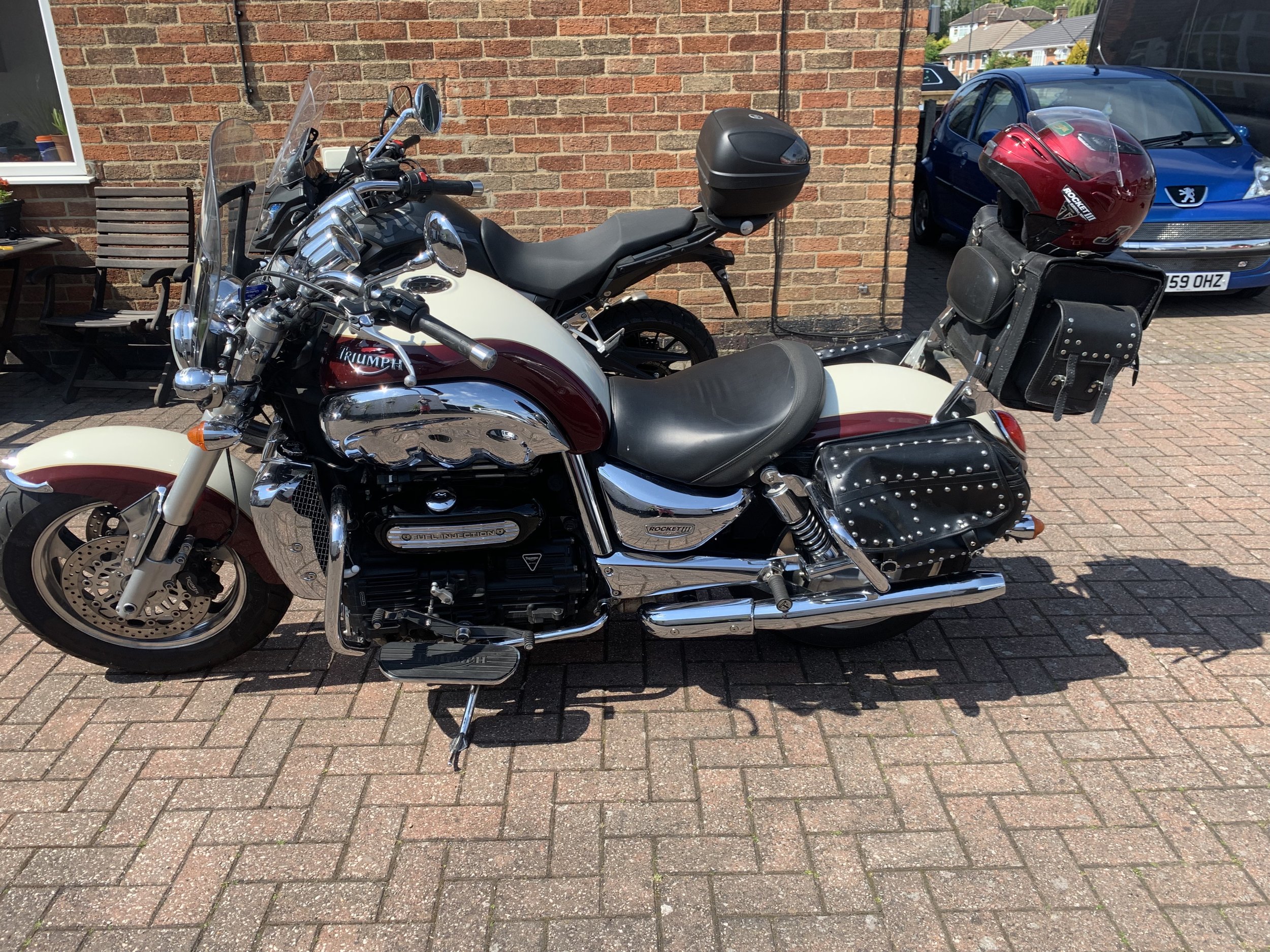 A Triumph motorcycle parked on a brick driveway, with a red and white helmet on a black leather saddlebag. In the background, there's a smaller motorcycle and a parked blue car, with a brick house wall behind.