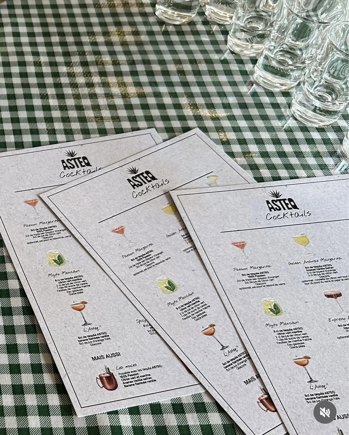 Menus de cocktails de l'établissement Asteq avec différentes recettes, posés sur une nappe à carreaux verts et blancs, avec verres shot